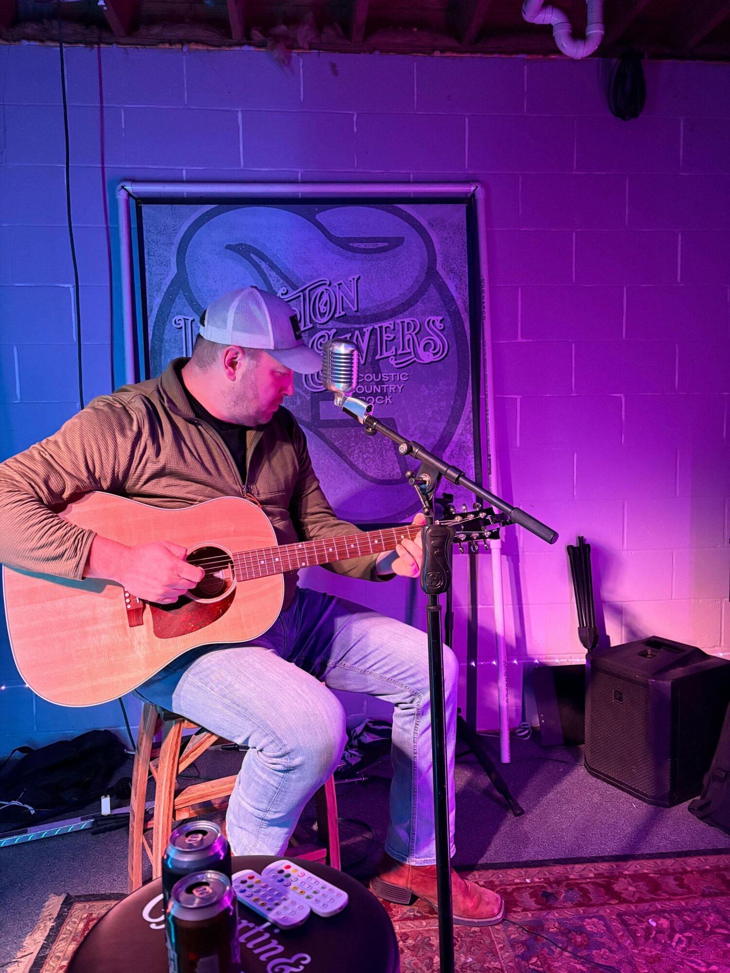 Person playing acoustic guitar on stage with purple lighting, microphone, and sound equipment nearby.