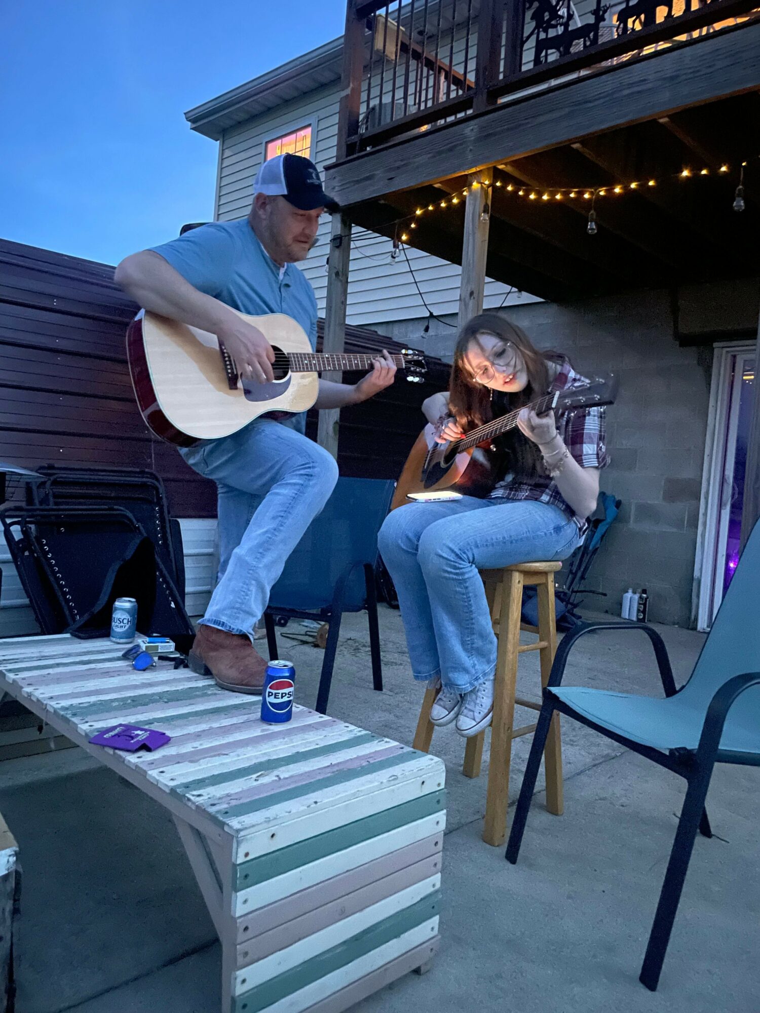 Two people playing guitar and singing on a rooftop with string lights, one sitting on a chair and the other on a box.