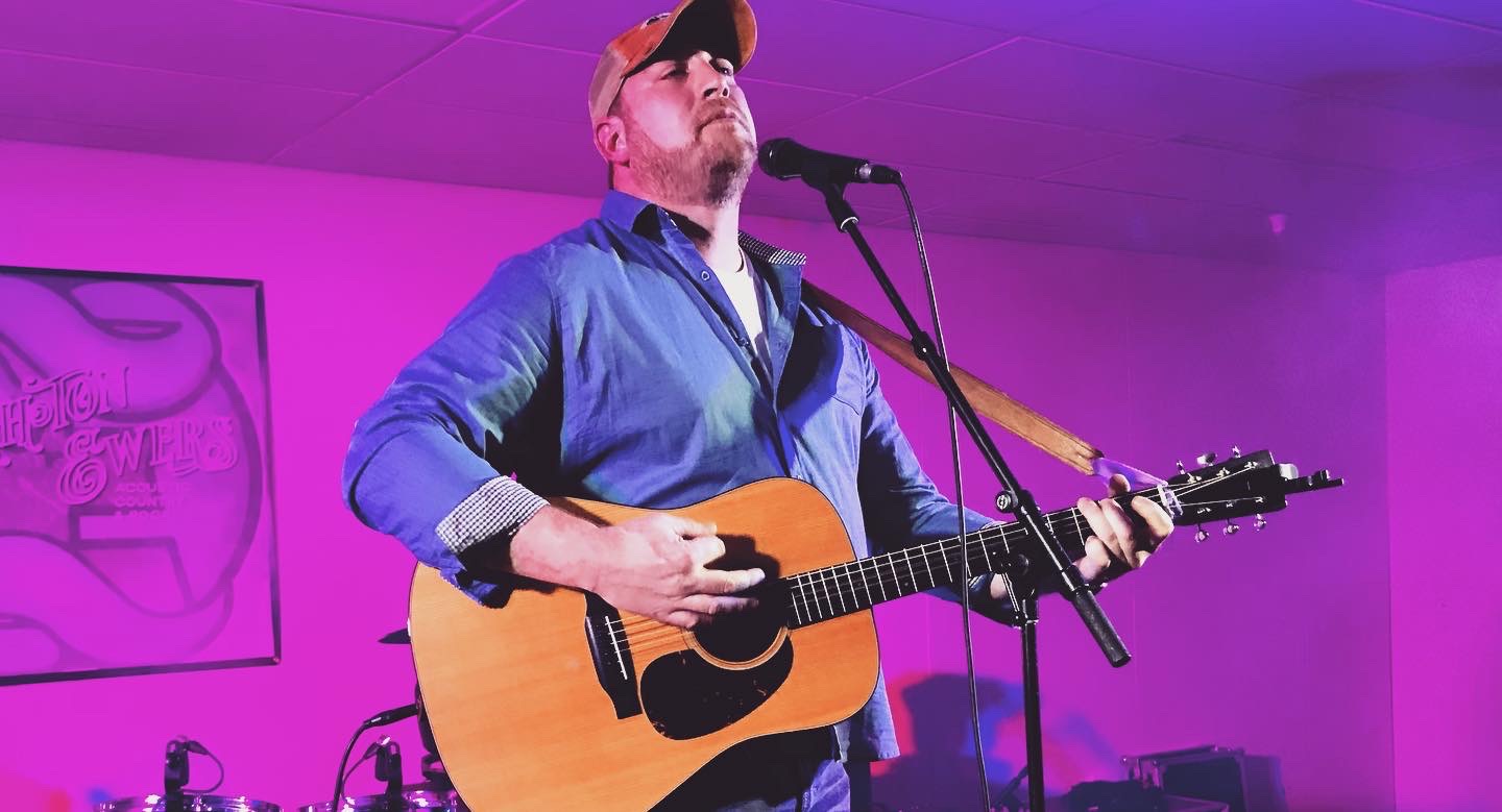 Person playing guitar and singing into a microphone on stage with purple lighting