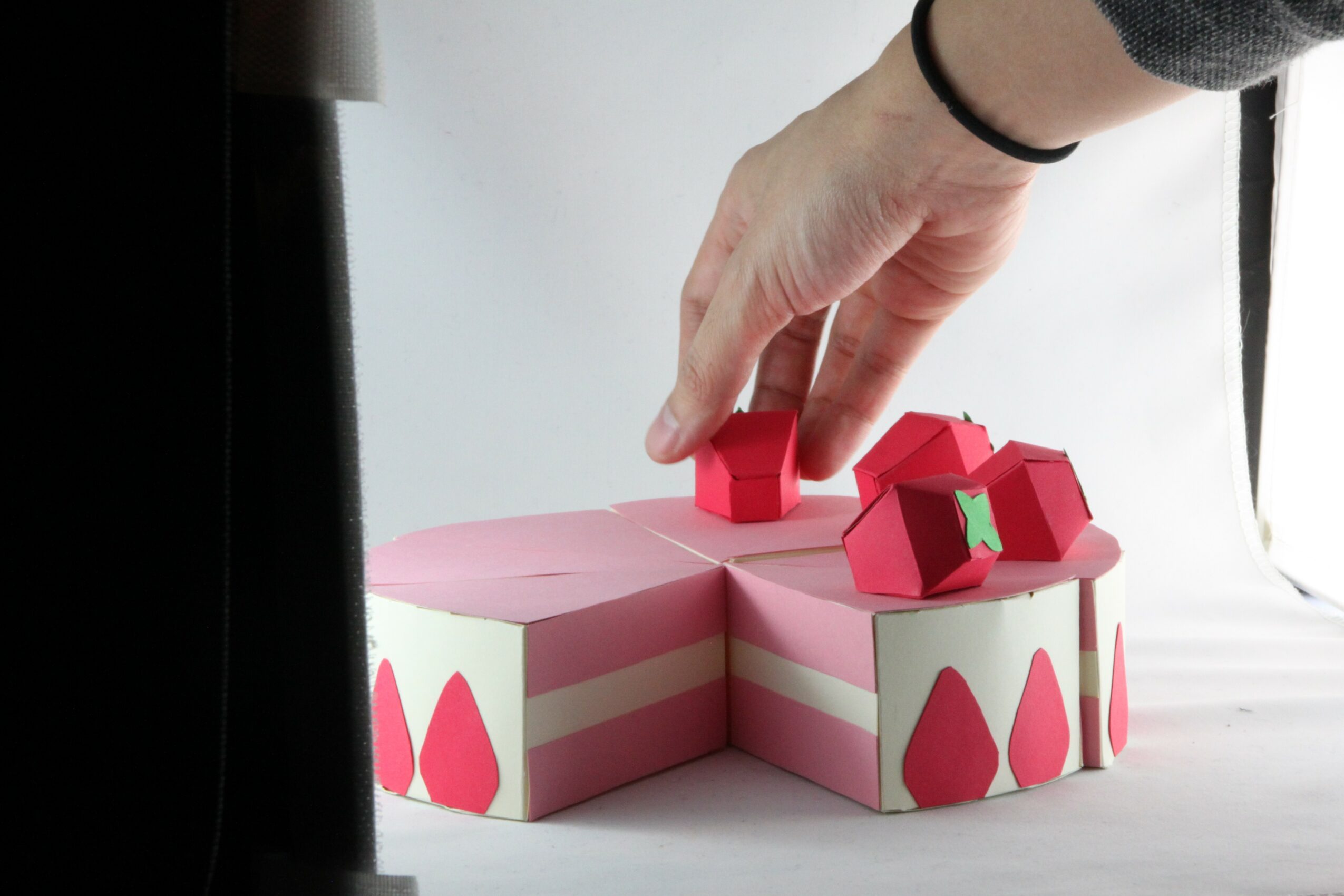 Hand placing a small red house-shaped object on a pink and white layered cake with teardrop decorations, against a white background.