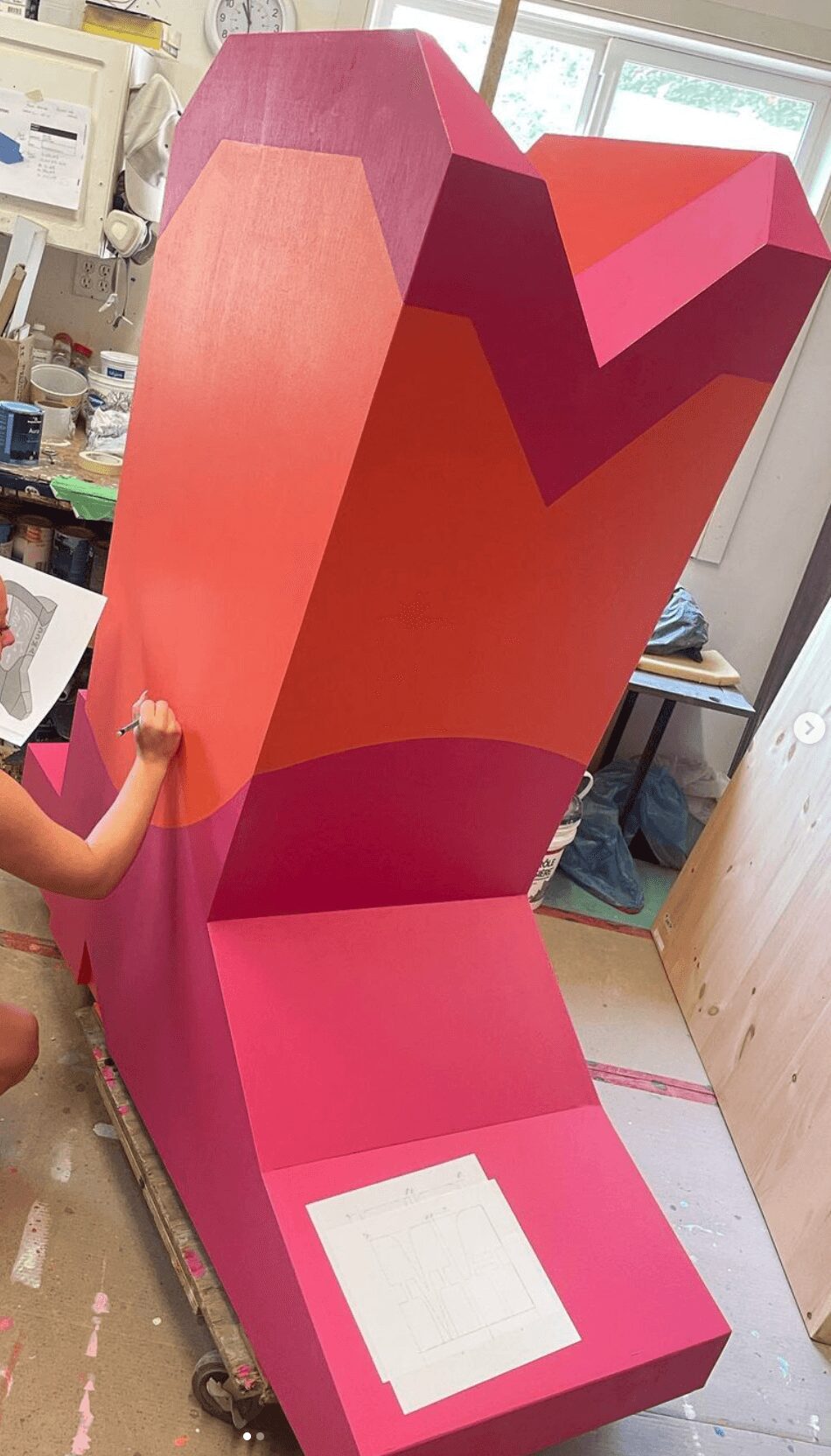 Large pink and purple heart-shaped structure in a room, with a child holding its side, and a window in the background.