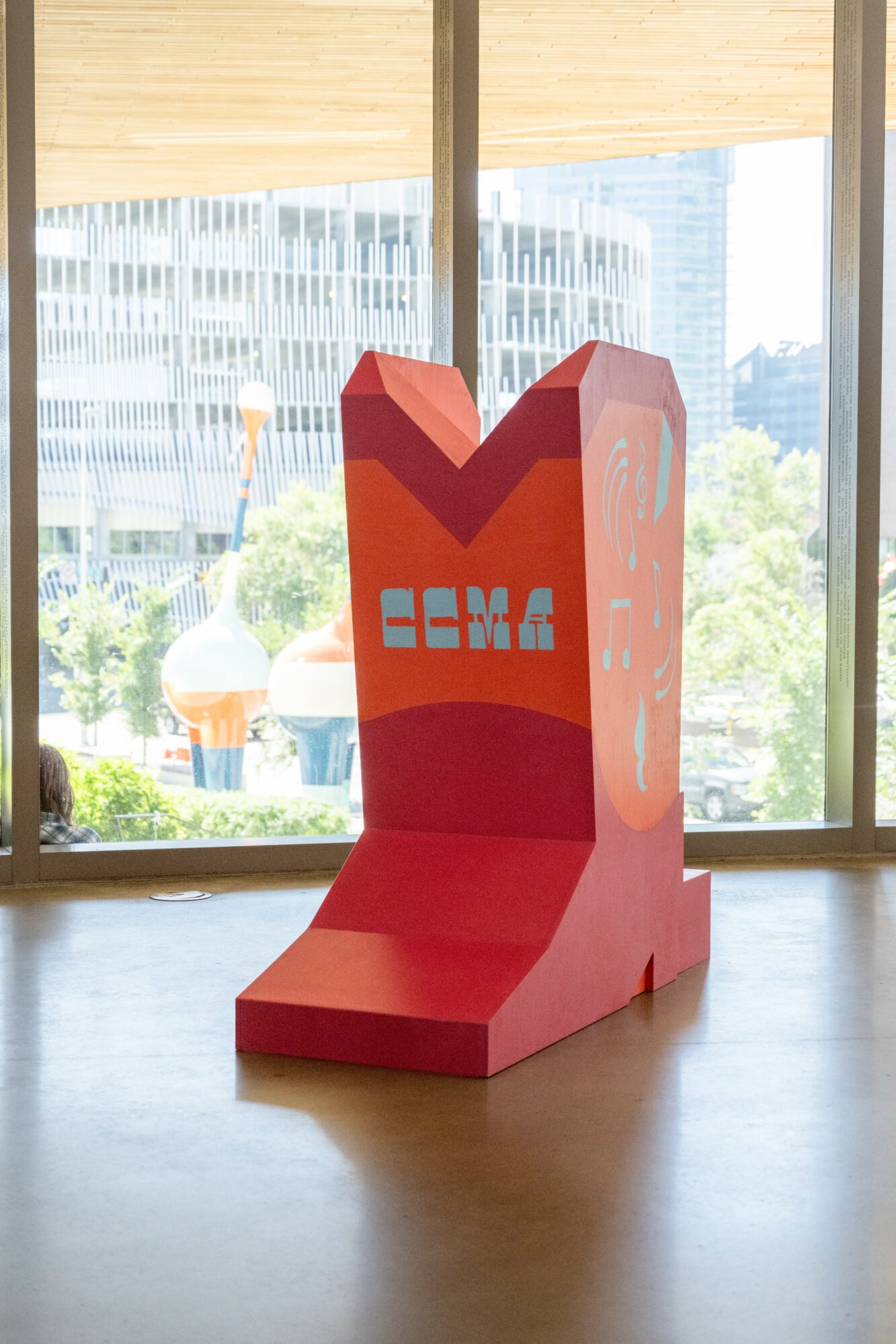 Red and orange sculpture with geometric shapes and text, placed indoors near large windows with cityscape view behind.