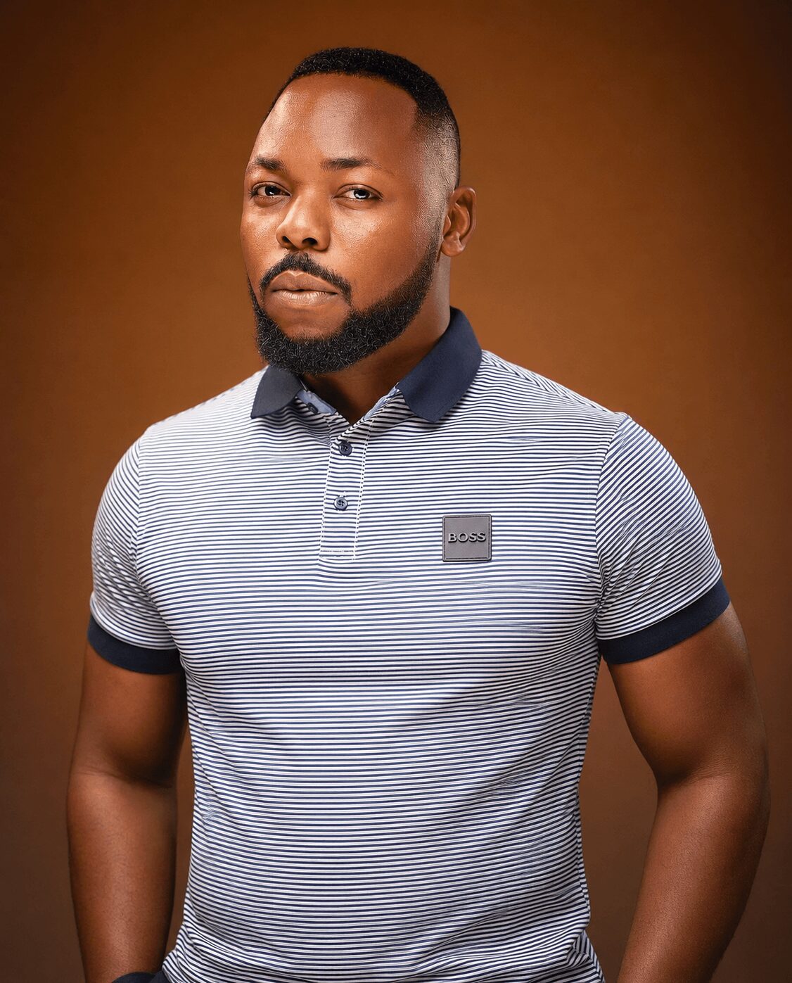 Man with a beard and short hair wearing a striped polo shirt, standing against a brown background.