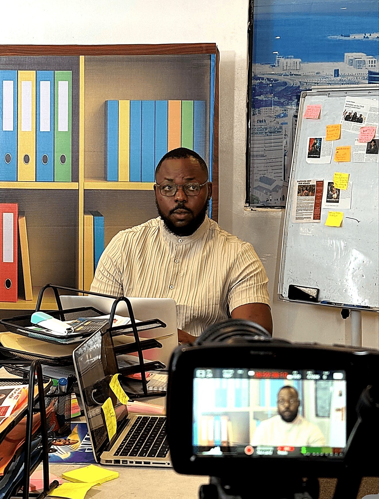 Man with glasses and beard sitting at desk in office, with bookshelf and whiteboard behind him.