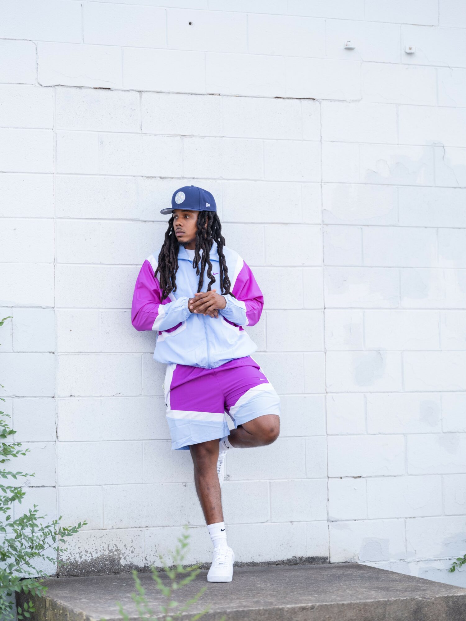 Person with long hair wearing a blue cap, purple and gray outfit, standing against a white wall.