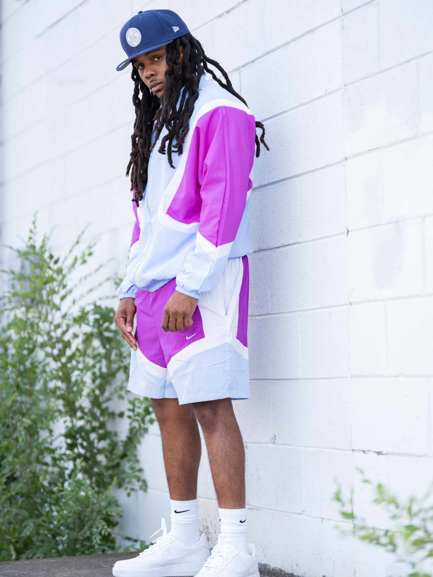 Young person with dreadlocks wearing a blue cap, pink and white hoodie, matching shorts, and white sneakers, standing against a white wall.