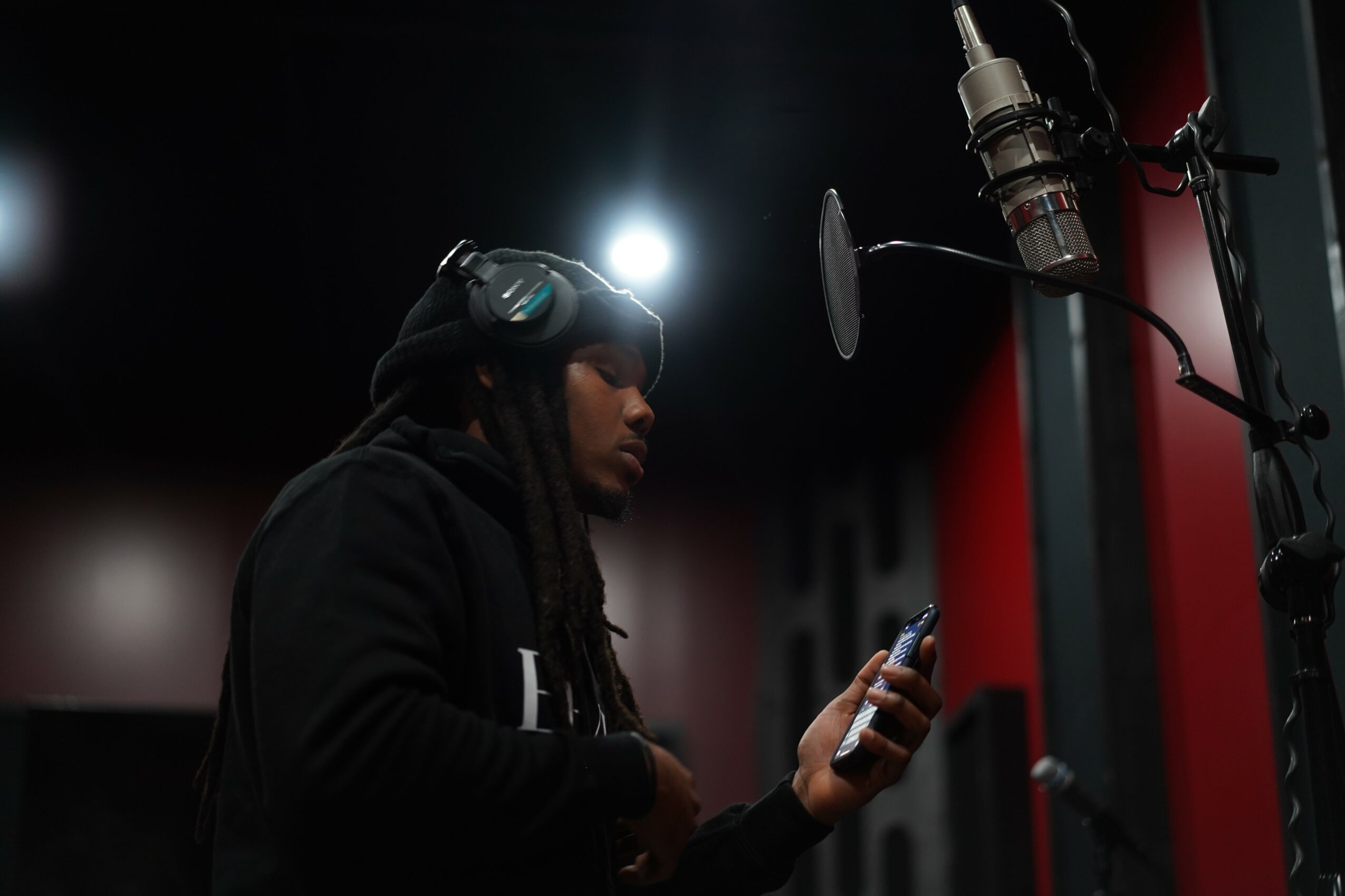 Person in a recording studio wearing headphones, holding a phone, with a microphone and soundproof walls visible.