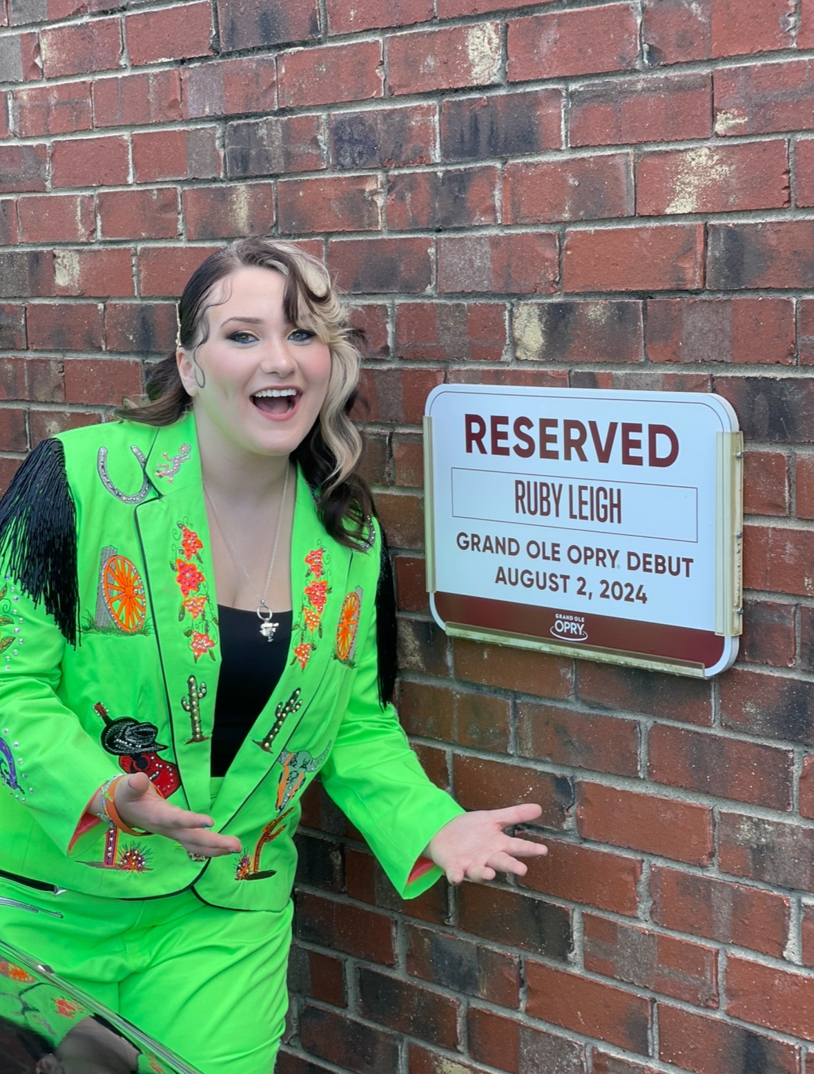 Woman in bright green outfit smiling and pointing at a reserved sign on a brick wall.