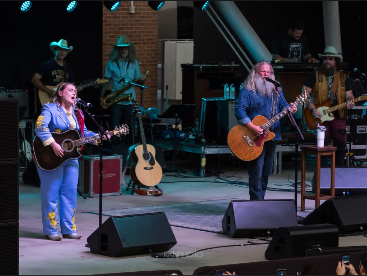 Band performs on stage with guitars, keyboard, and drums, under blue stage lights, brick wall background, and musical equipment.