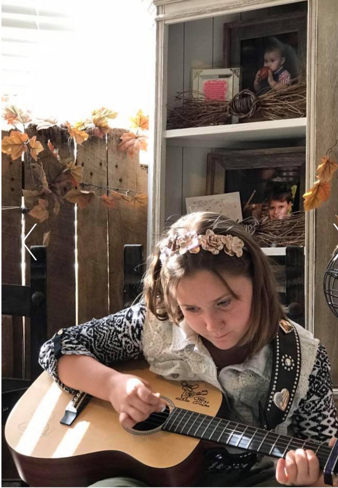 Girl with a floral headband playing an acoustic guitar indoors, with a cabinet and autumn decorations in the background.