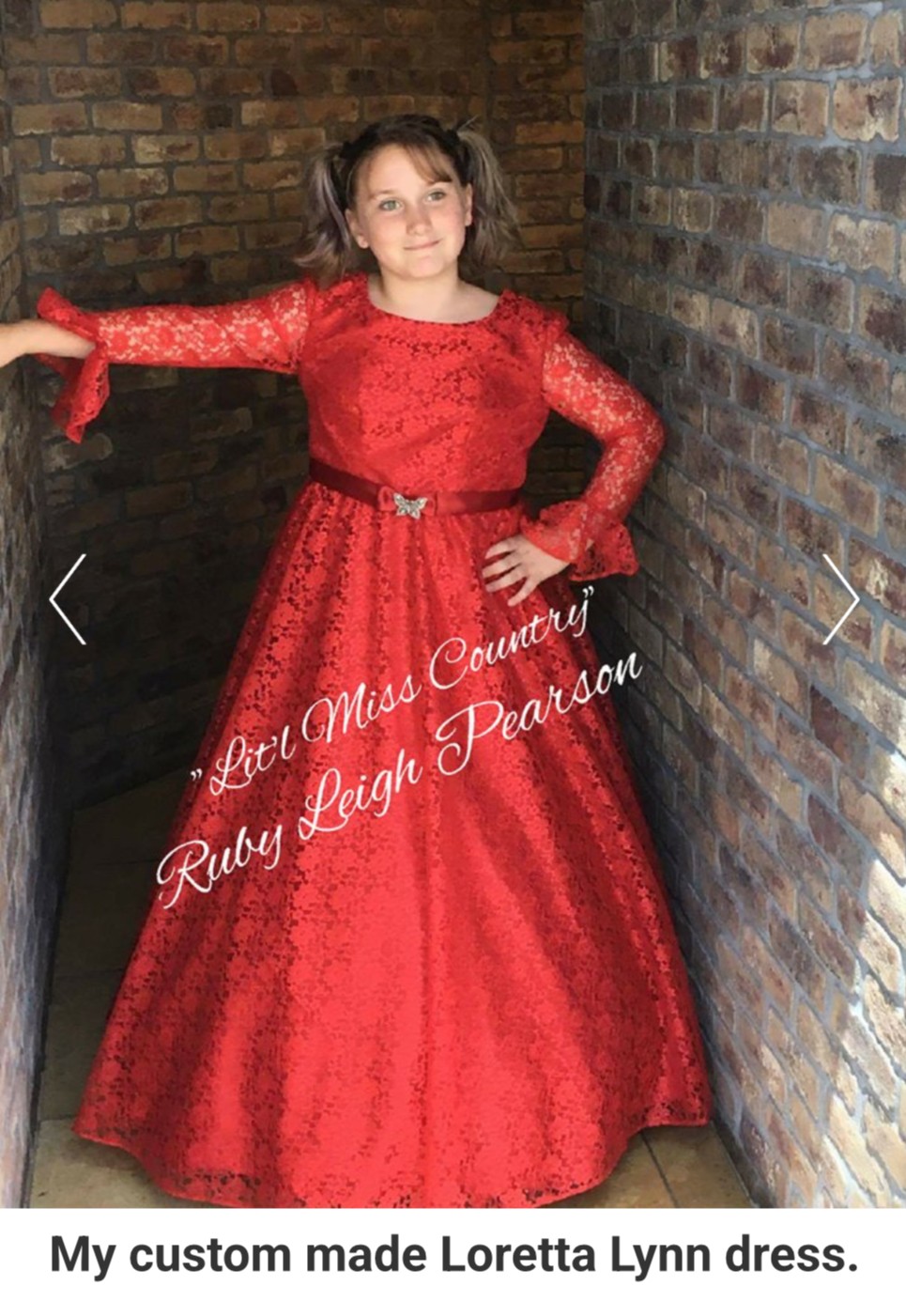 Young girl in a red lace dress with long sleeves and a full skirt, standing against brick walls, smiling.