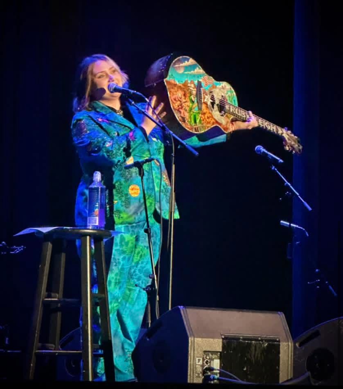 Performer singing into a microphone on stage with a decorated guitar hanging above her, wearing a colorful outfit, with a stool and sound equipment nearby.