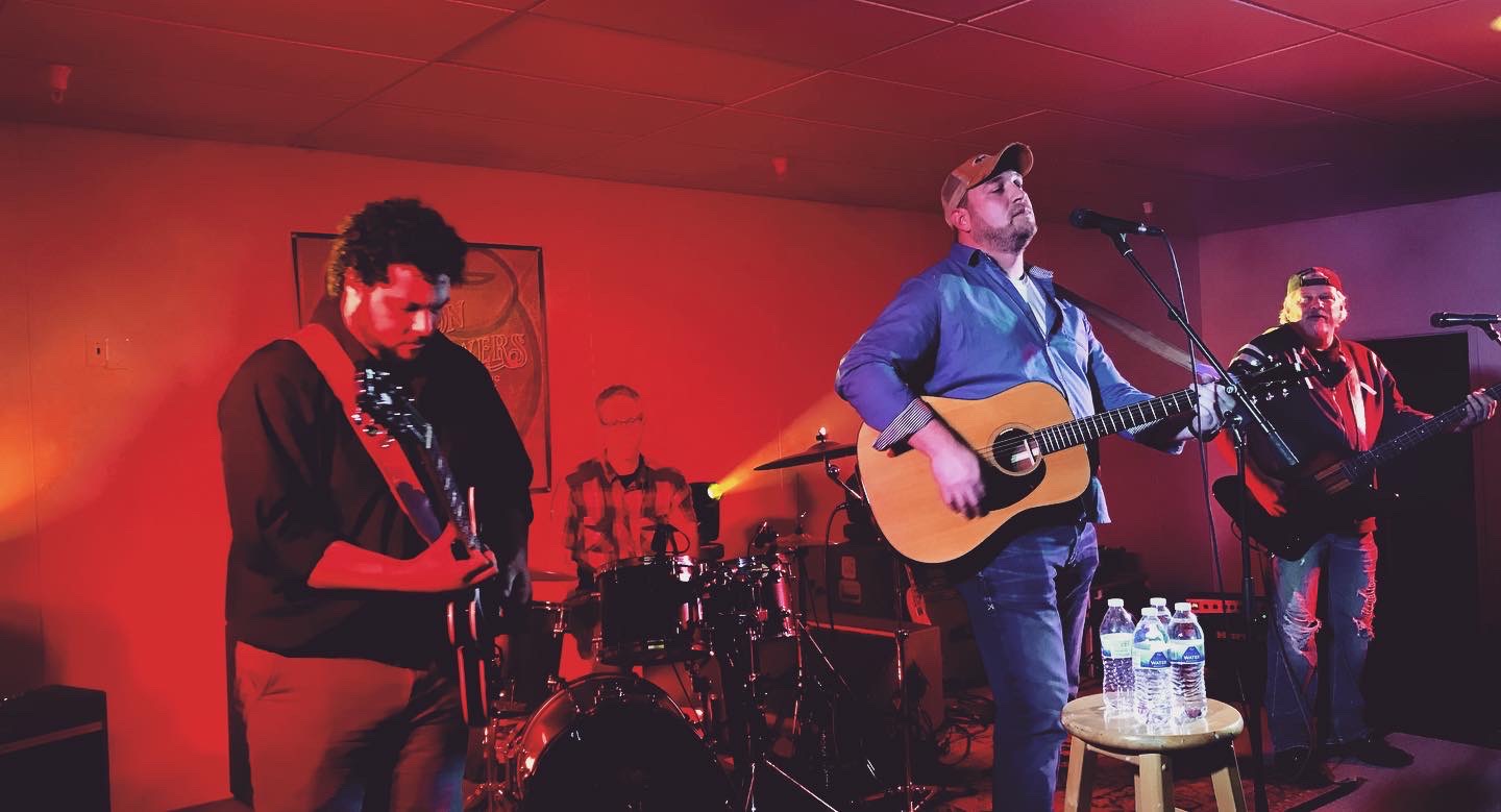 Band performs on stage with four members, one playing guitar and singing, others playing instruments, in a dimly lit room.