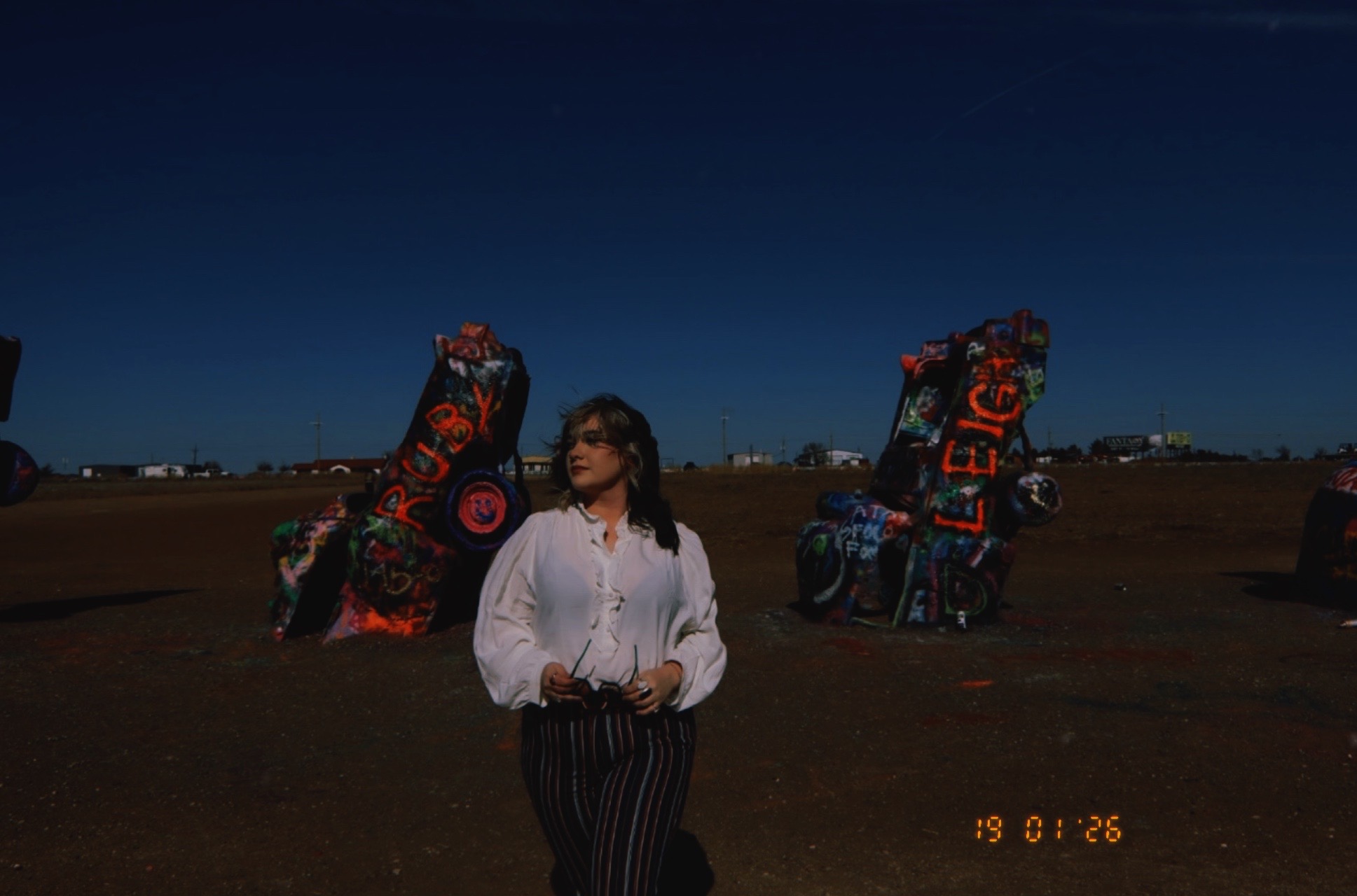 Woman standing outdoors with two large decorated objects behind her, clear blue sky, and a flat landscape.