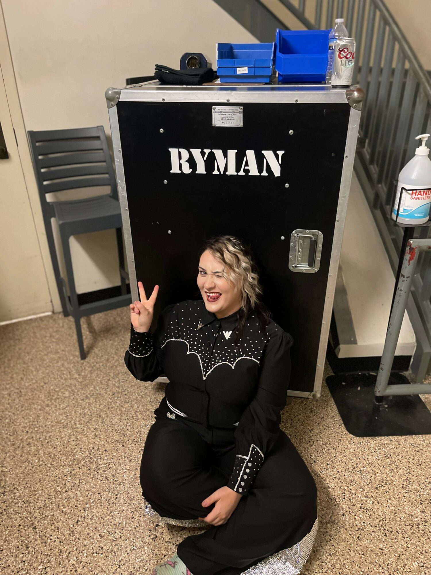 Person sitting on the floor, smiling, making a peace sign, in front of a black RYMAN cabinet, with a chair and sanitizer nearby.