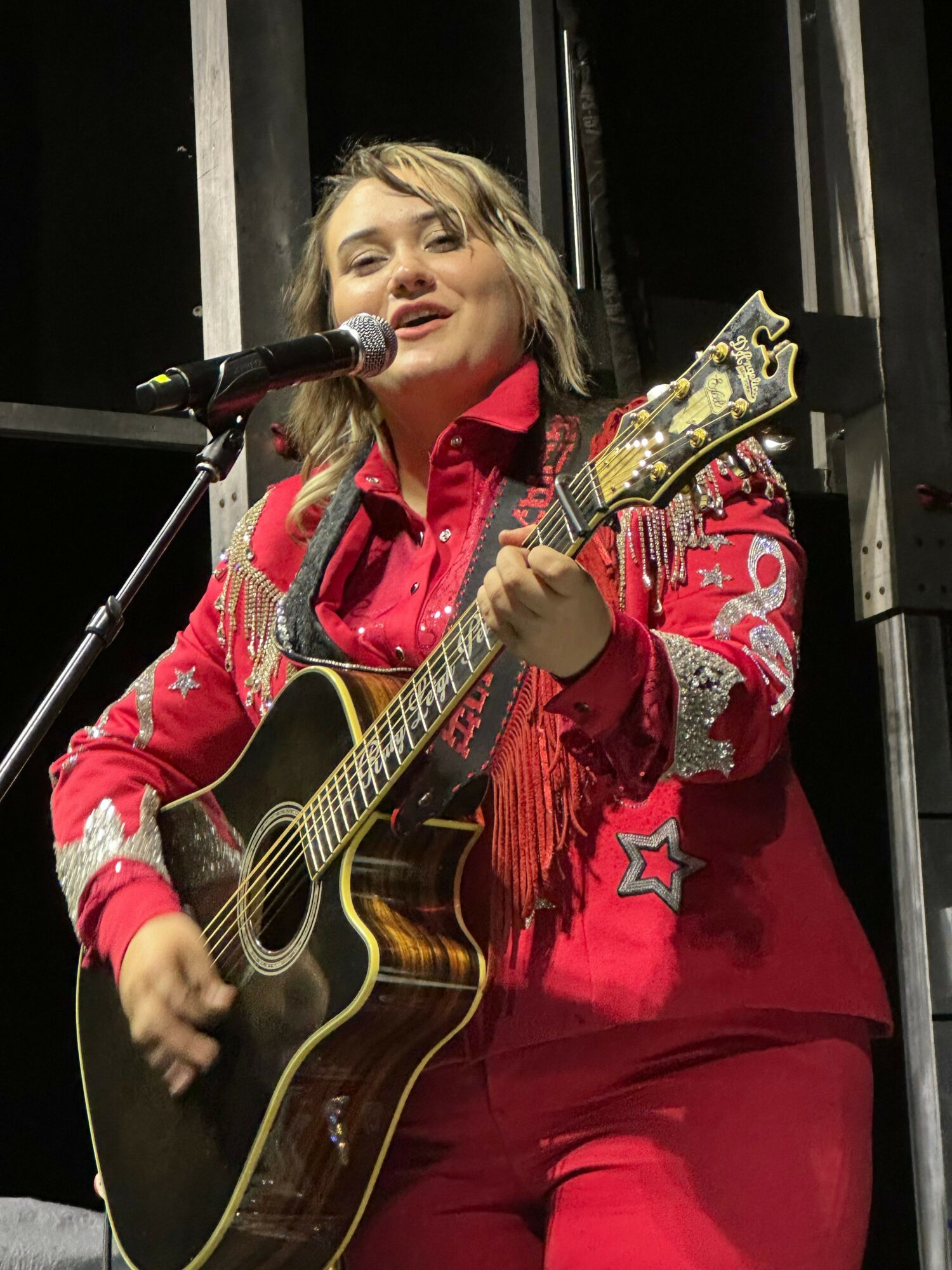 Person singing into a microphone while playing an acoustic guitar, wearing a red jacket with star patterns, on stage.