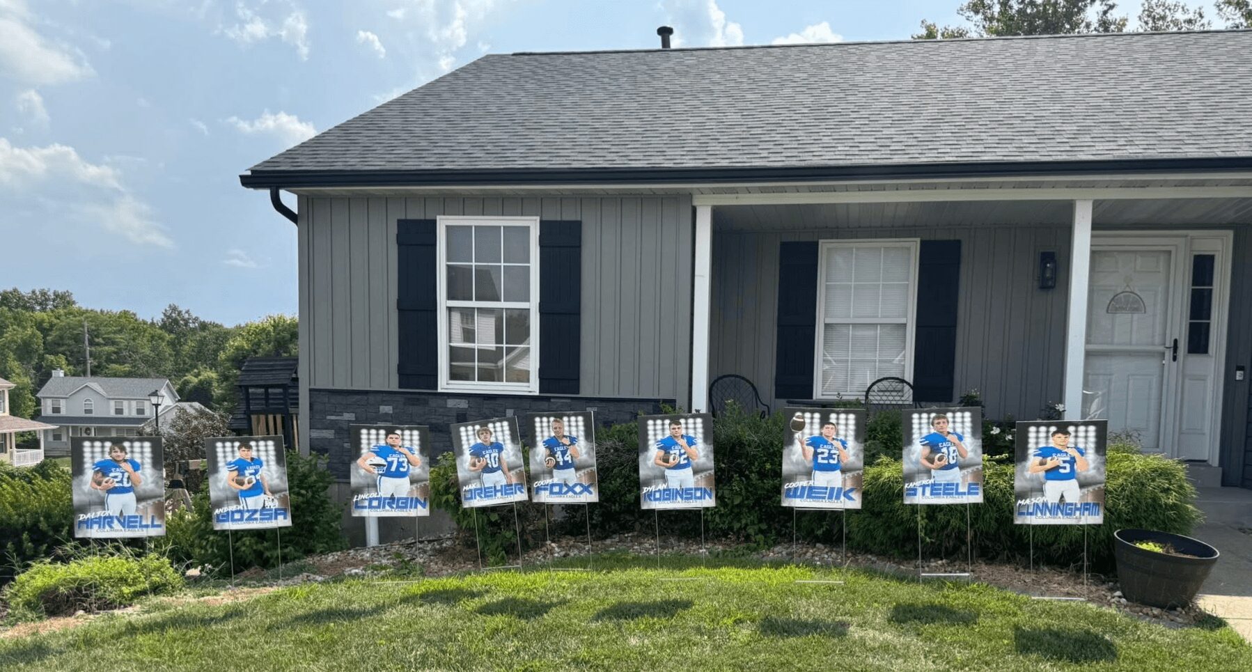 House with a row of eight election campaign signs in front yard, black fence, gray siding, and black shutters.