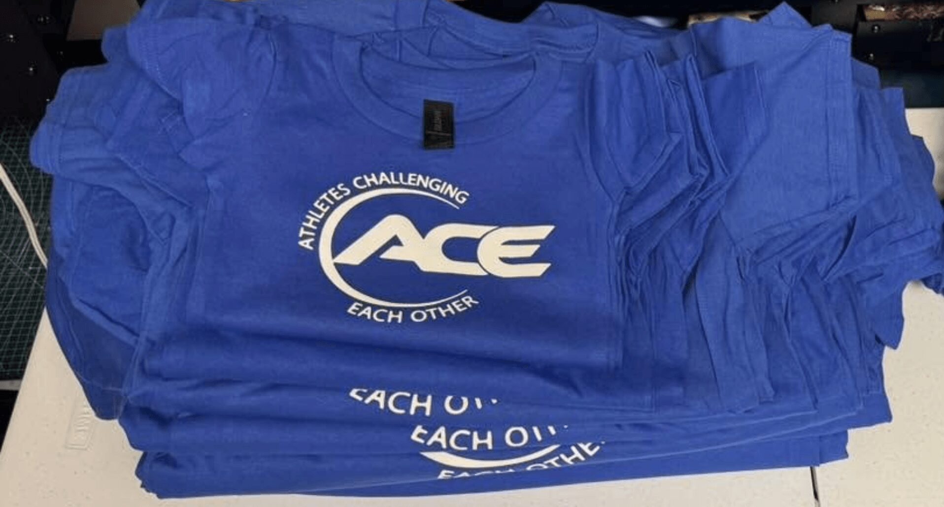 Stack of blue T-shirts with white logo and text, ruffled sleeves, on a dark surface.