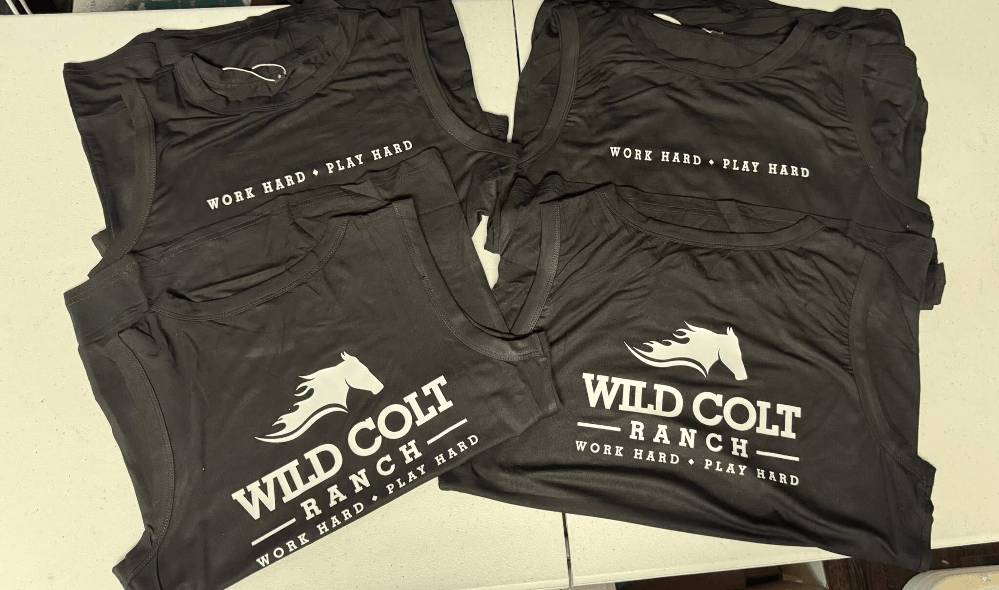 Four black sports shirts with white logo and text, folded and stacked, on a light surface.