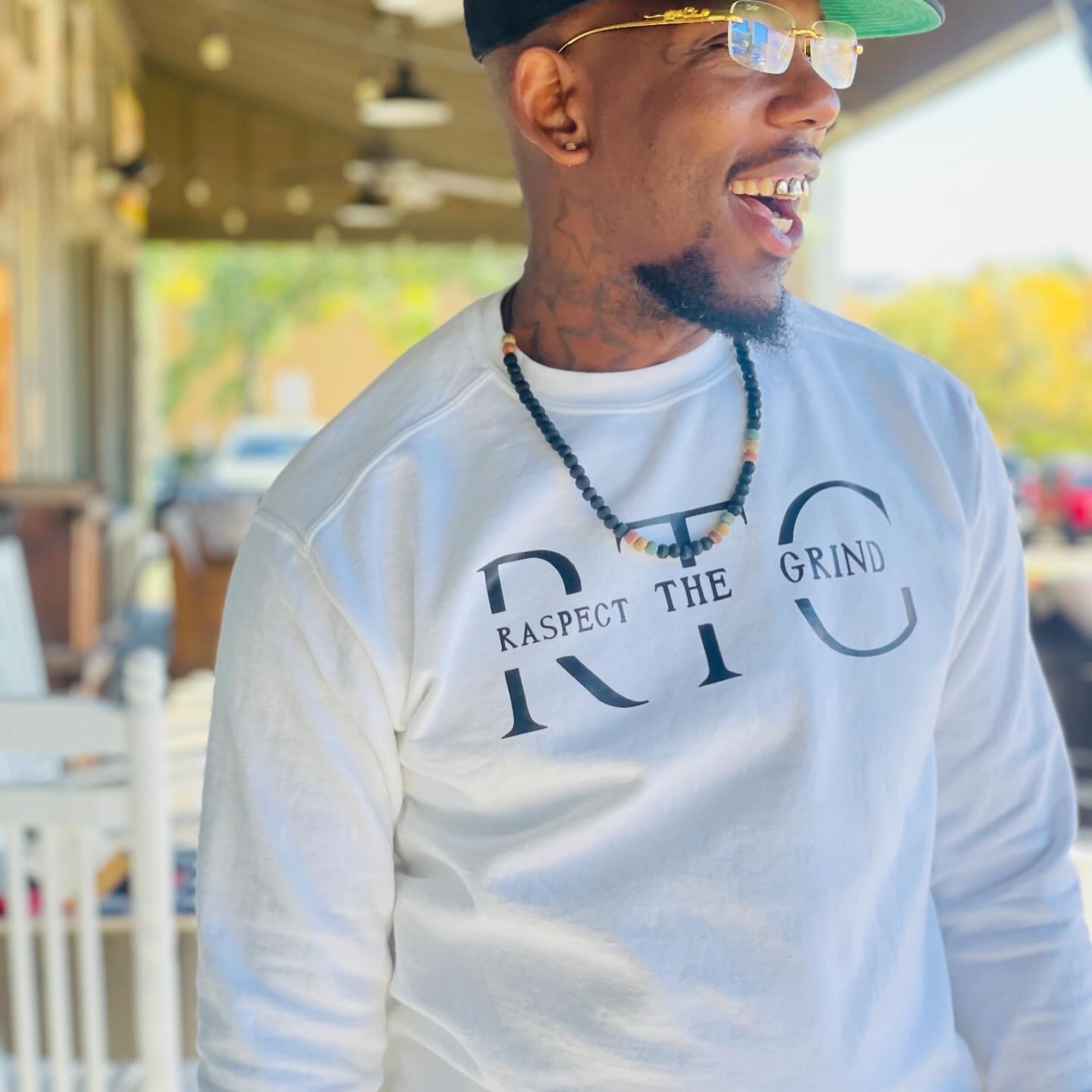 Man wearing glasses, a cap, and a white sweatshirt outdoors, smiling and looking to the side.
