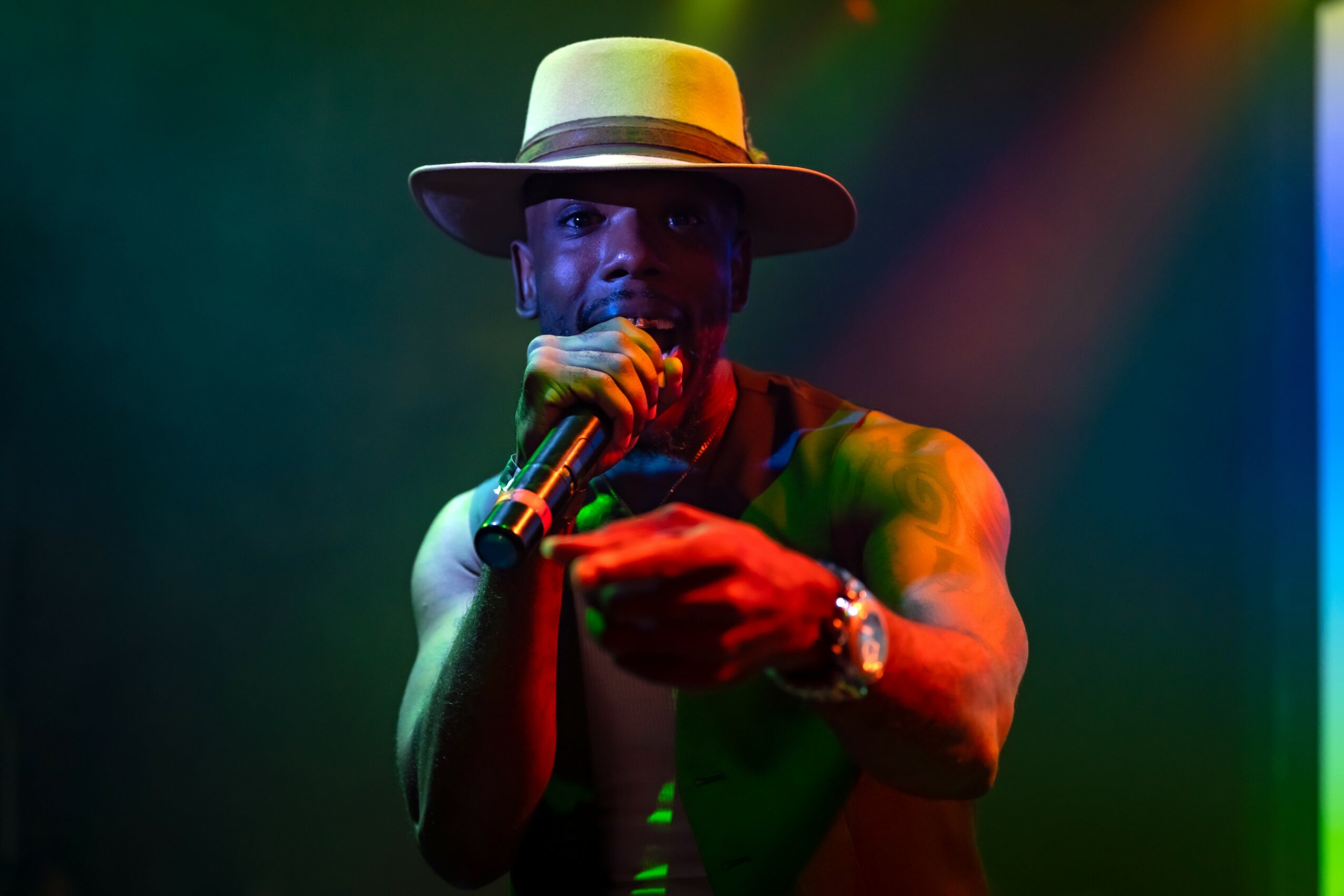 Person wearing a wide-brimmed hat, holding a microphone, on stage with colorful lighting, dark background.