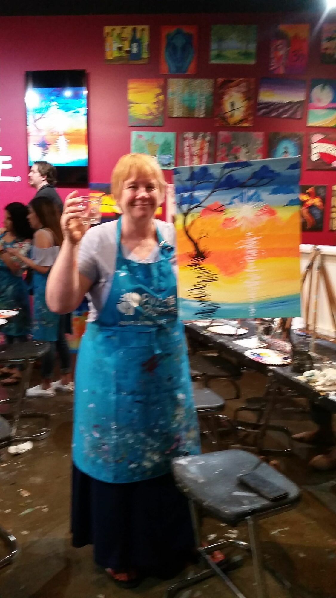 Woman in blue dress standing in art studio with colorful paintings on wall behind her.