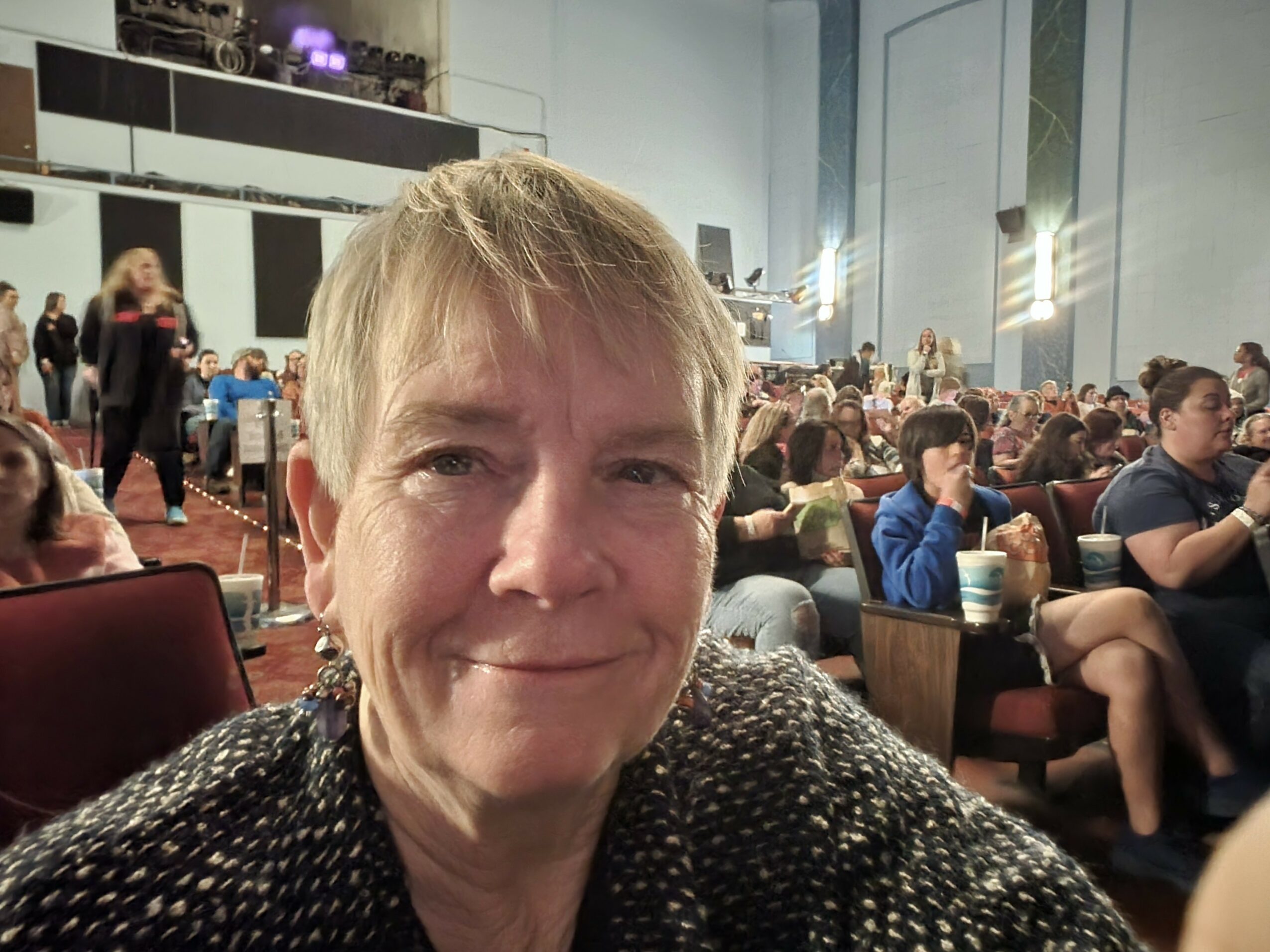 A woman with short blonde hair smiling in a large indoor event with many seated people and a stage in the background.
