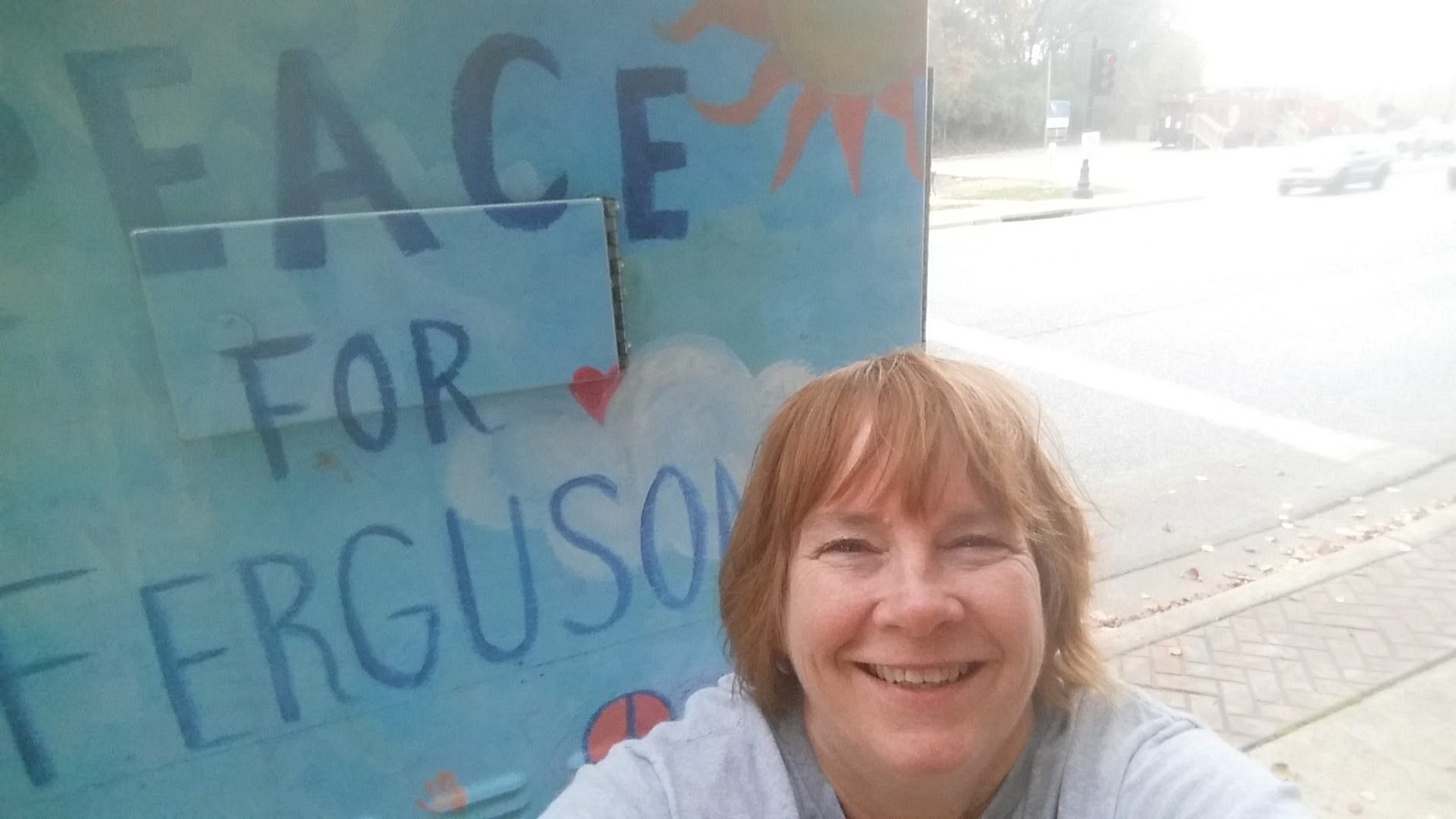 Smiling woman with short hair in front of a colorful sign that reads 'PEACE FOR FERGUSON'.