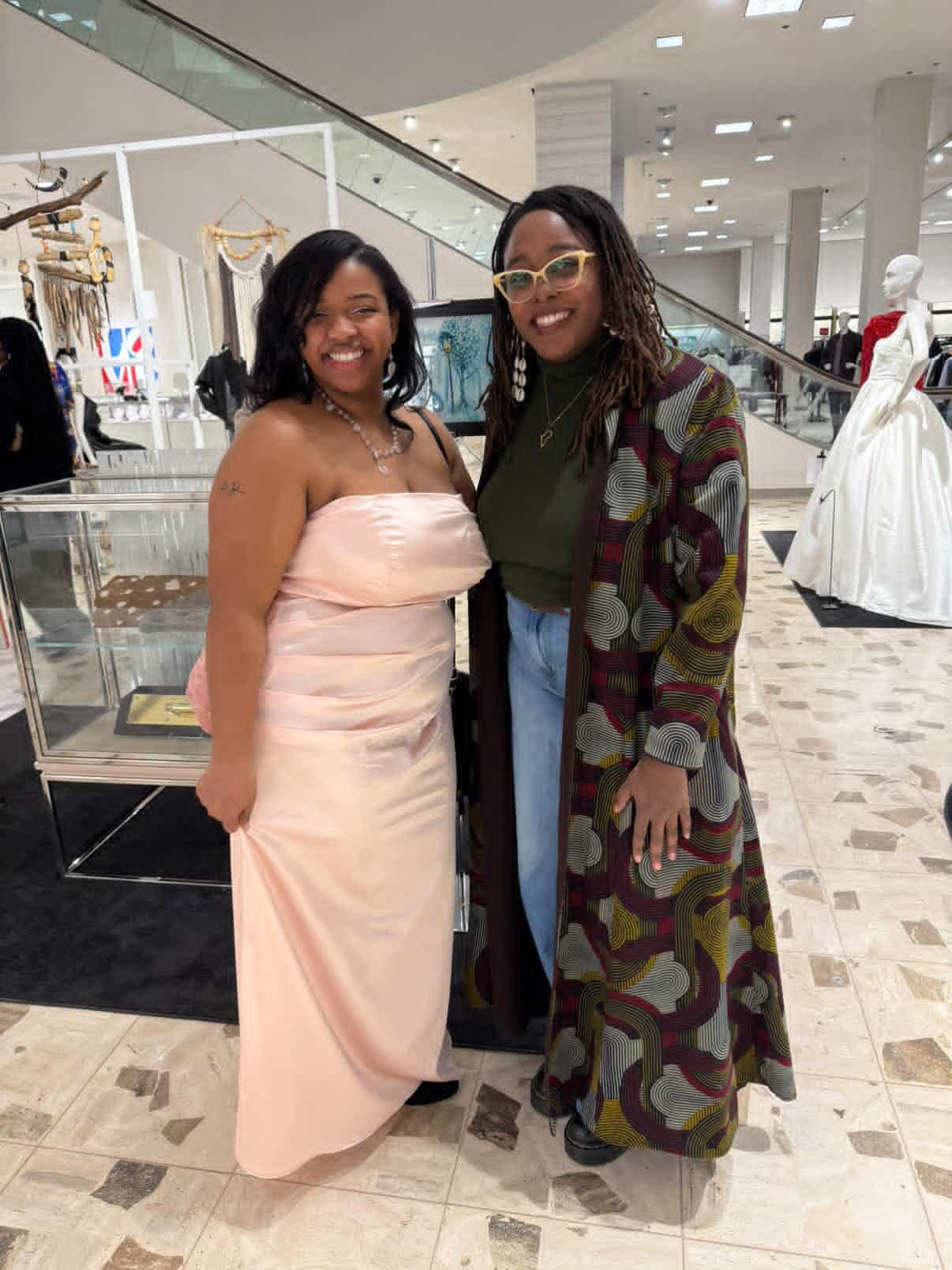 Two women standing inside a shopping mall, smiling, with clothing displays and mannequins in the background.