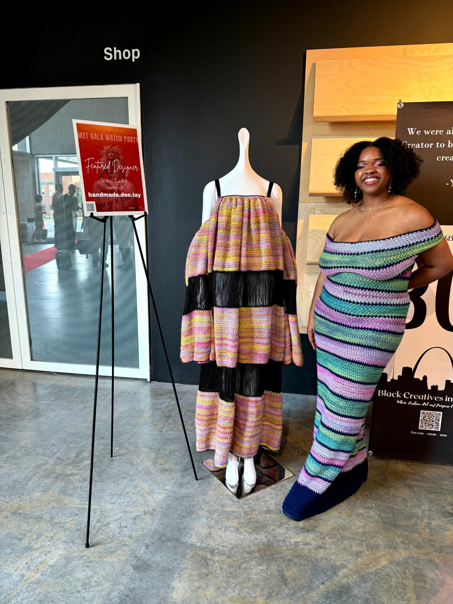 Woman with curly hair in a colorful striped off-shoulder dress standing next to a mannequin wearing a multicolored dress, in a store setting.