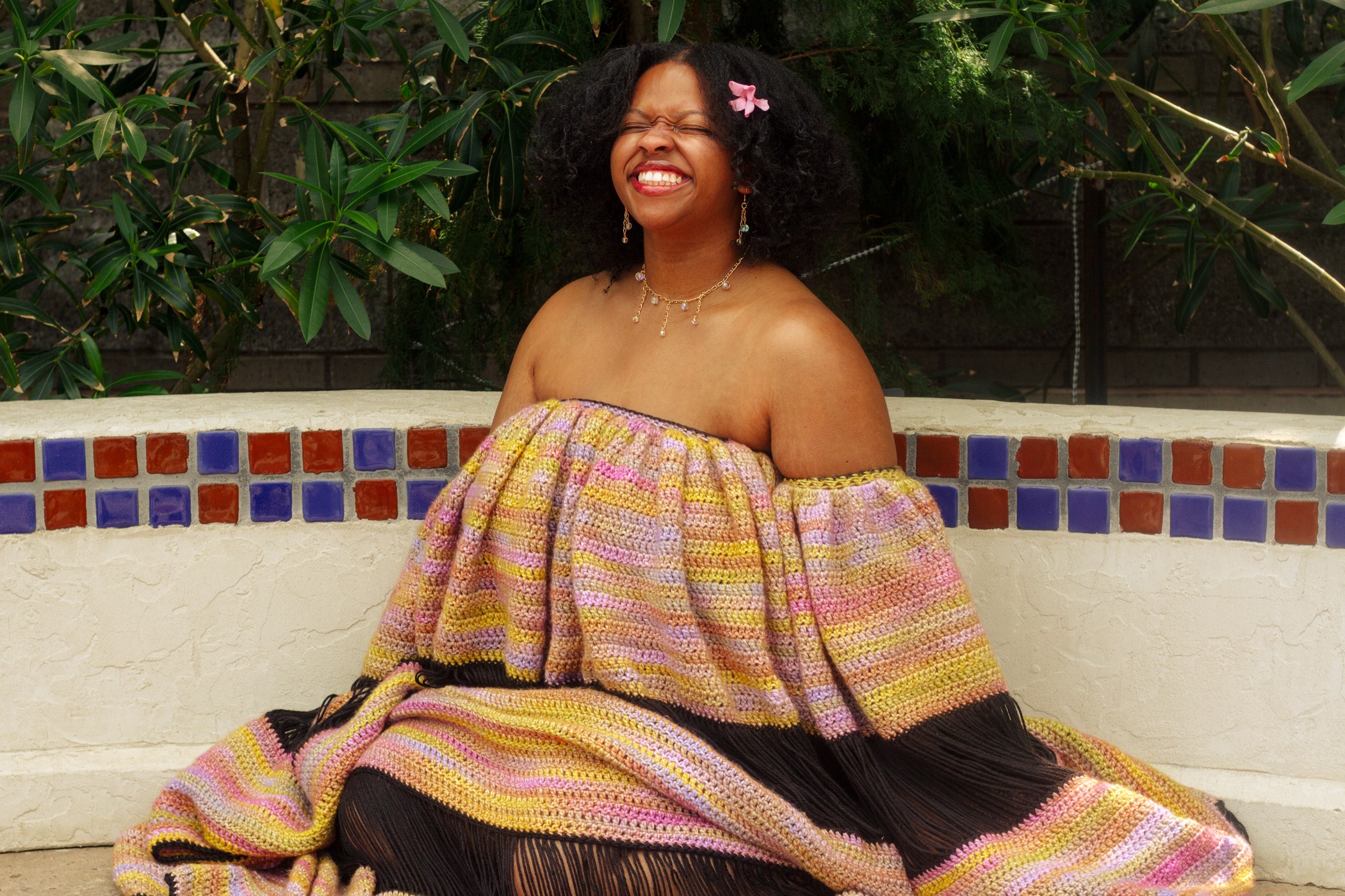Smiling woman with curly hair and a pink hair clip, wearing an off-shoulder multicolored dress, sitting outdoors near a tiled wall.