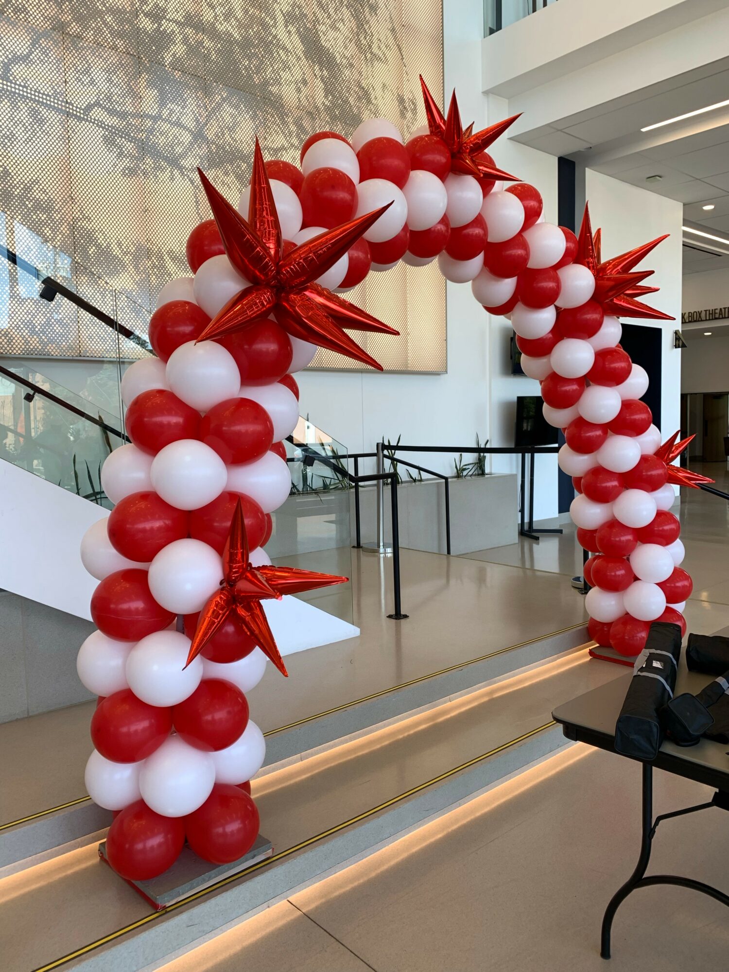 Arch made of red and white balloons with star-shaped decorations, set indoors near stairs and large windows.
