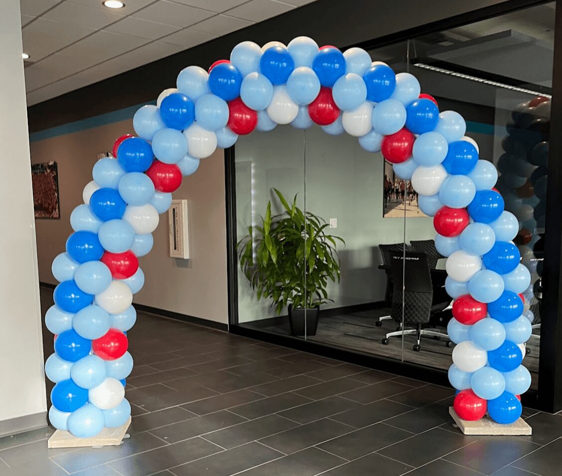 Balloon arch with blue, white, and red balloons in front of a glass wall with a plant inside an office.