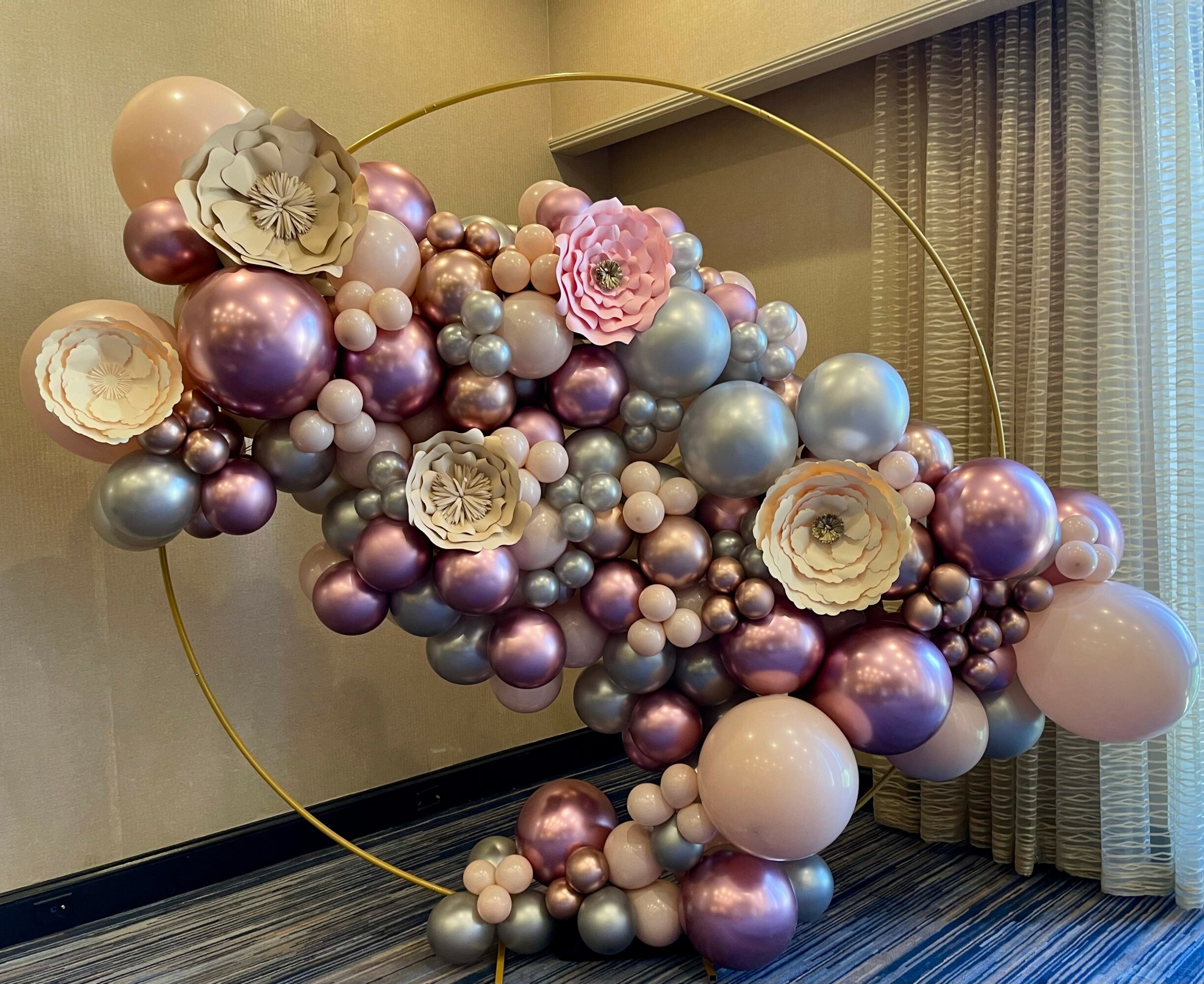 Decorative arrangement of pink, purple, and silver balloons with large paper flowers, inside a circular frame, on a wall.