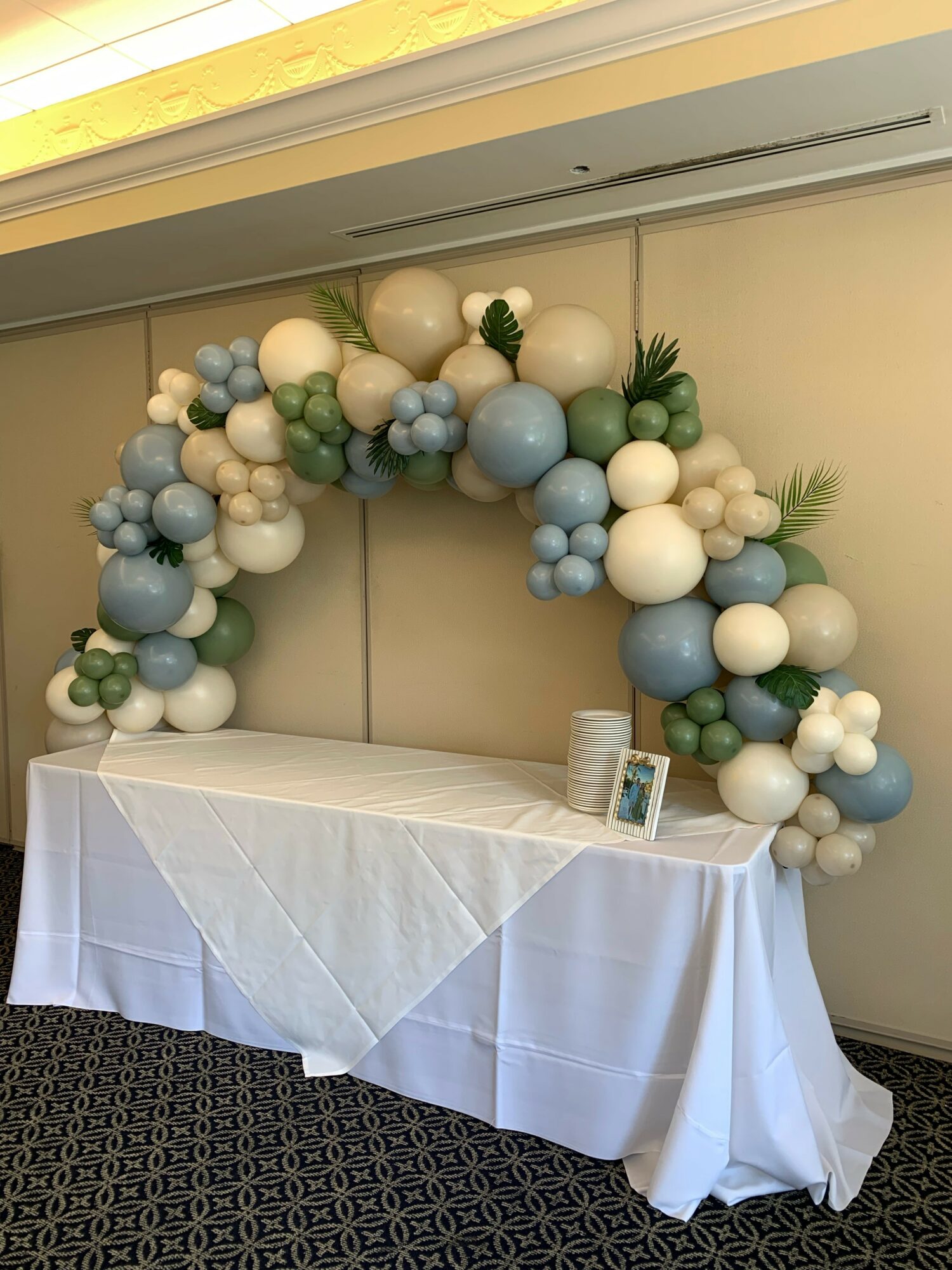 Balloon arch with white, blue, and green balloons over a table with a white tablecloth, a small framed picture, and a cup.