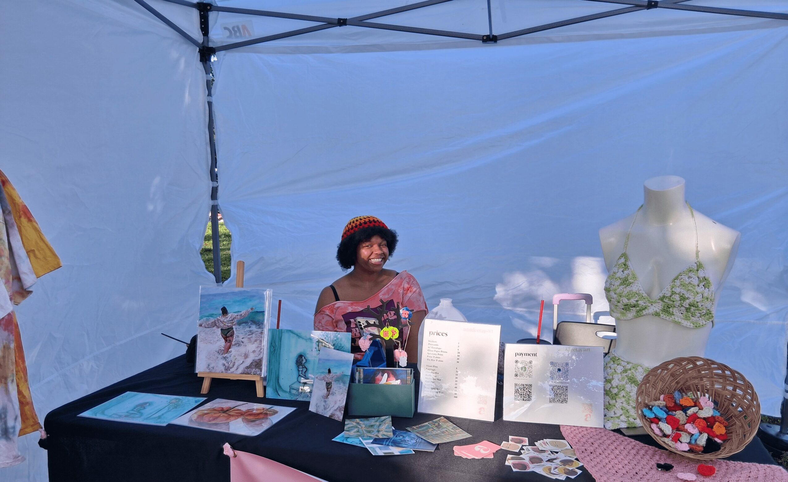 Woman standing behind a table with various items inside a blue tent, mannequin with green floral design, and colorful objects.