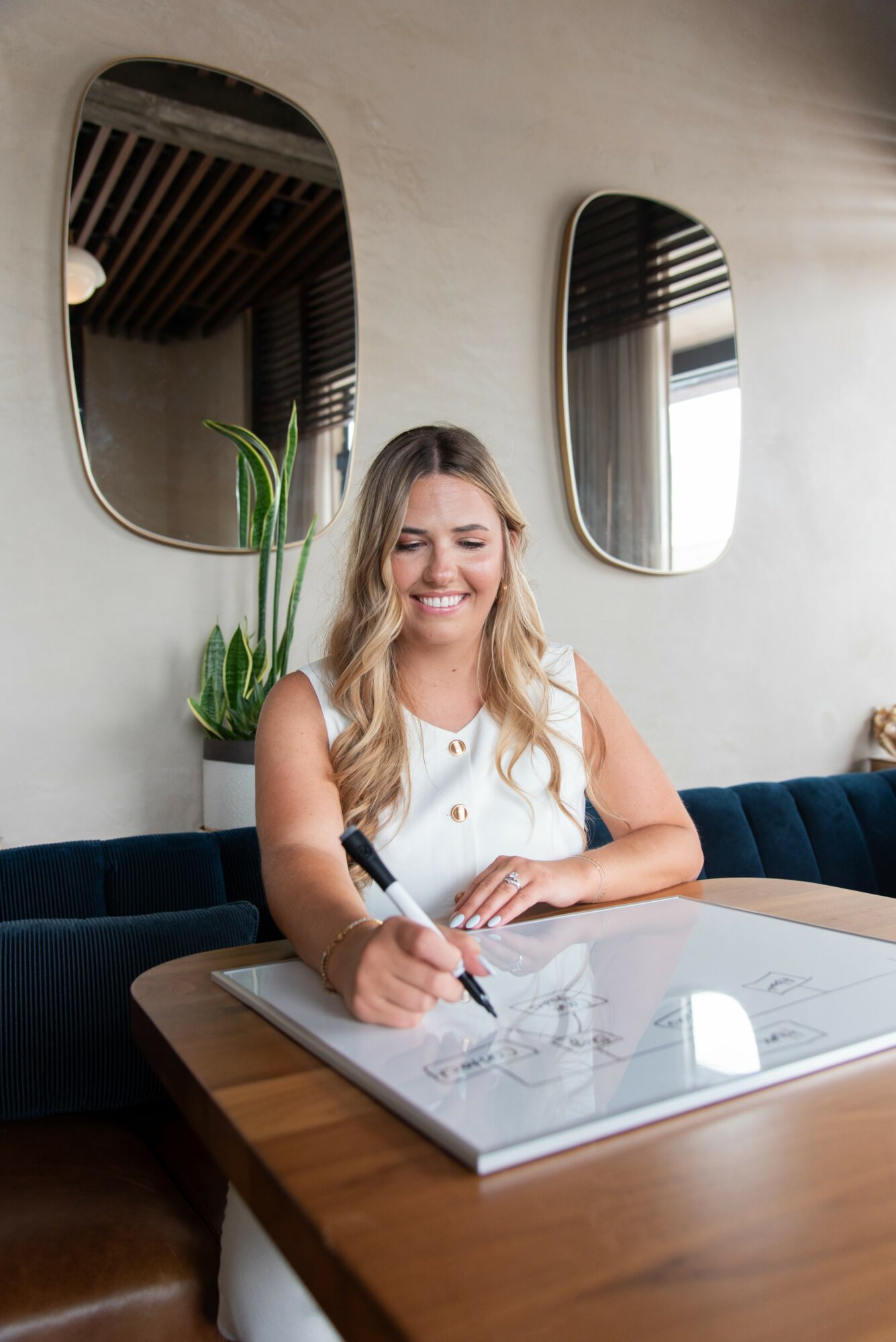 Woman with long blonde hair smiling while writing on a whiteboard at a table, with mirrors and a plant in the background.