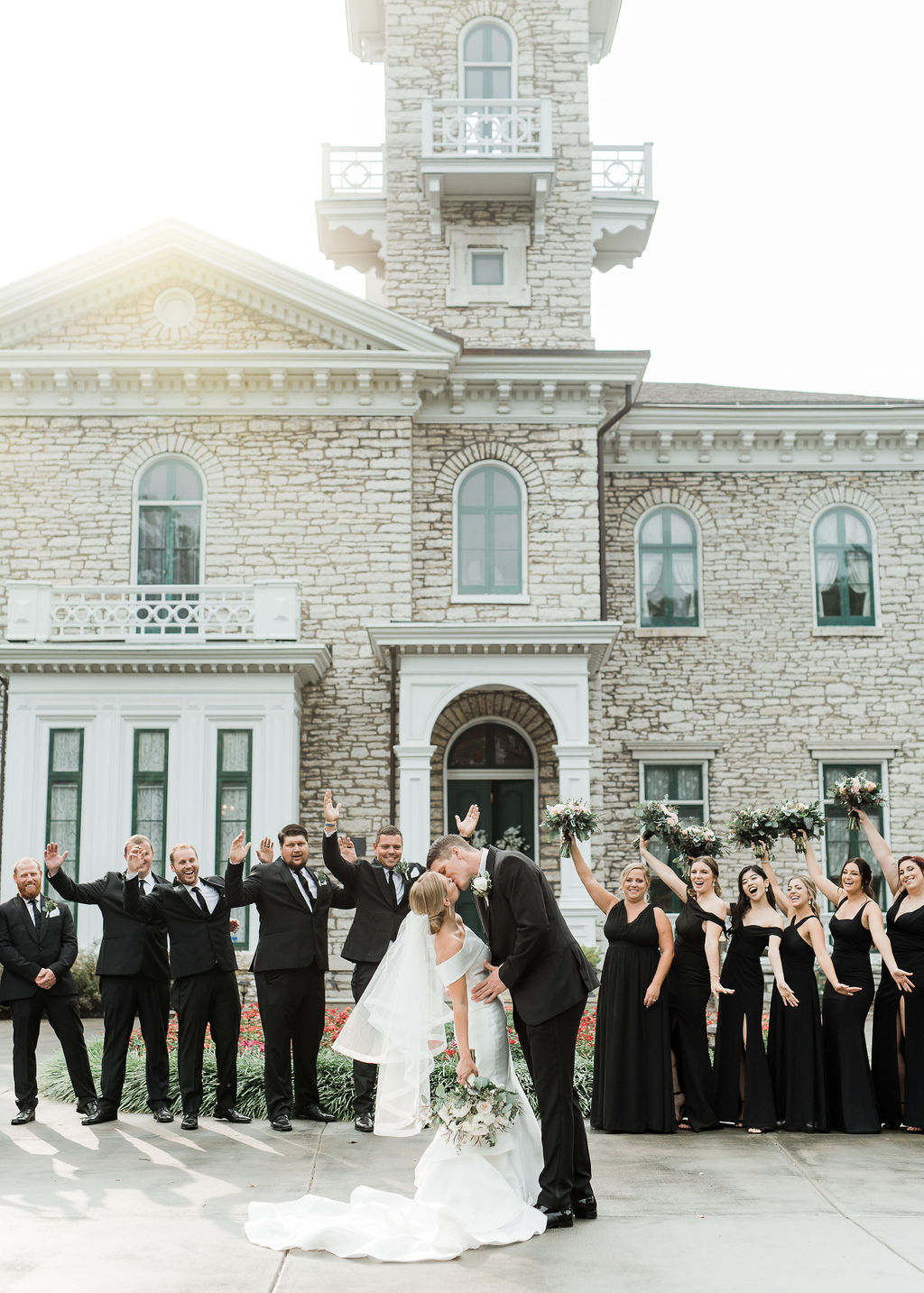 Bride and groom kissing in front of a historic stone building with wedding guests in formal attire, some waving hands.