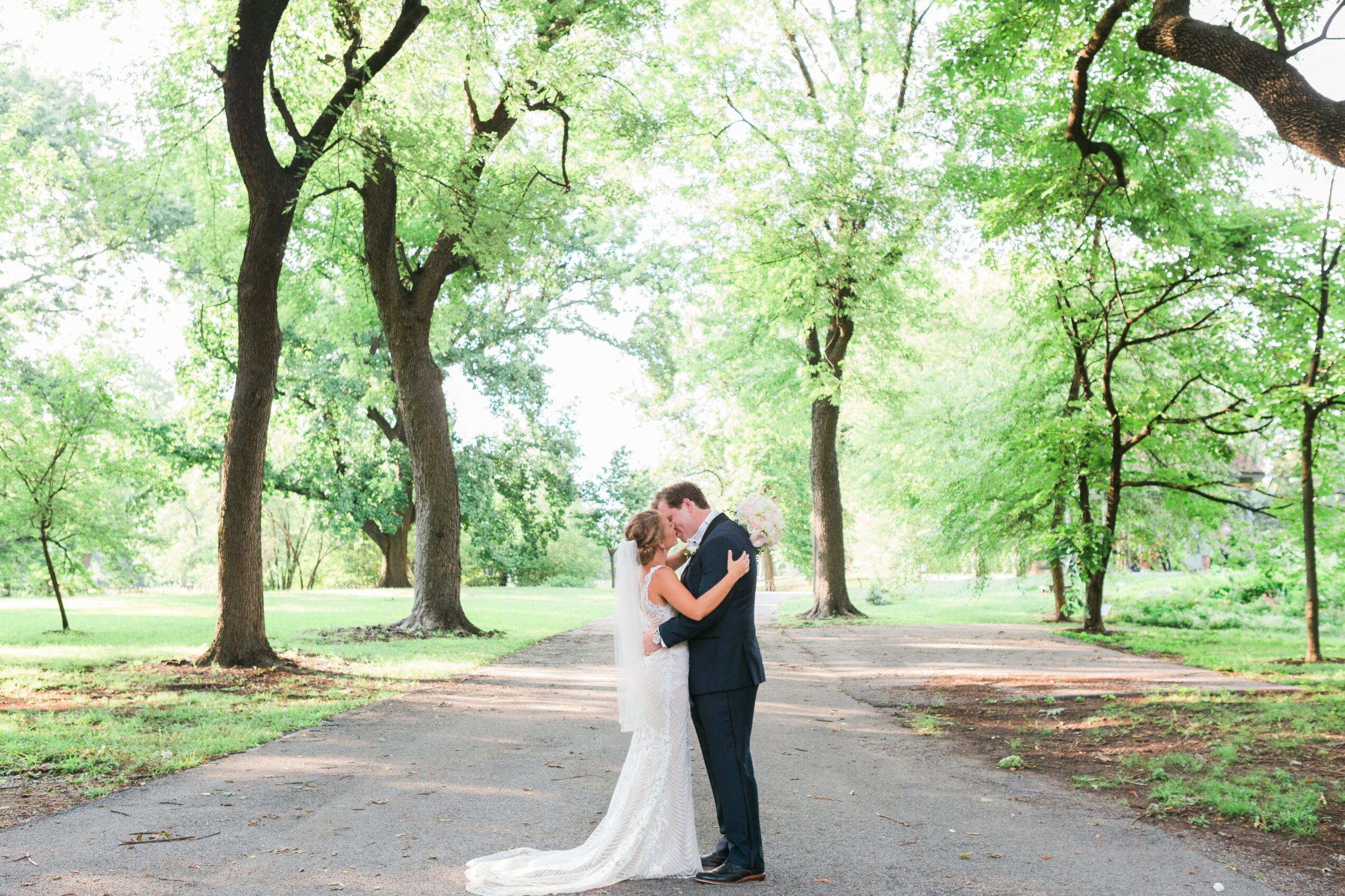 A couple in wedding attire standing on a tree-lined path, embracing and looking at each other, surrounded by green trees.