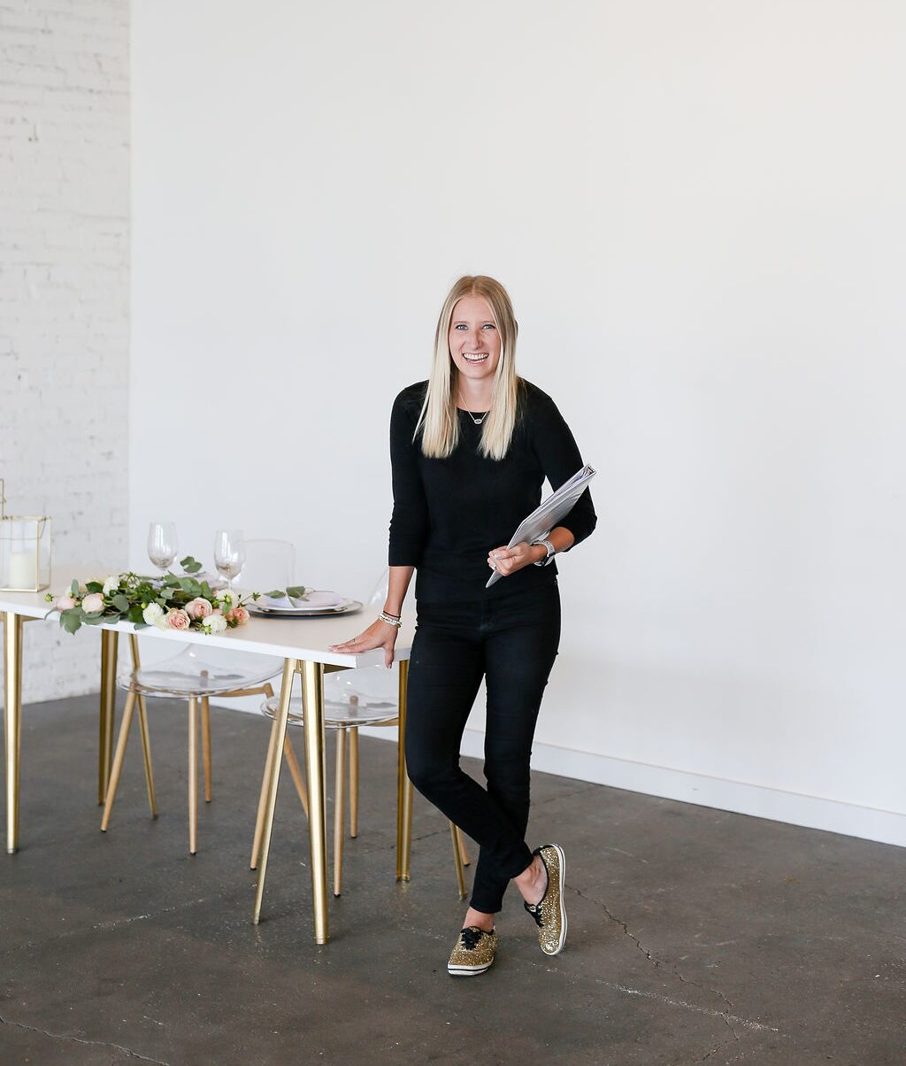 Woman standing next to a table with flowers and glasses, holding a notebook, smiling, in a bright room.