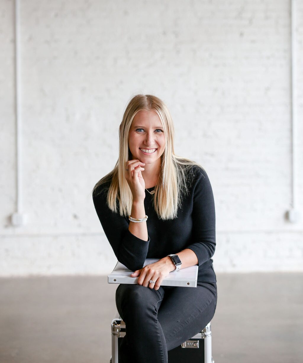 Young woman with long blonde hair, smiling, sitting on a stool in a bright, minimalistic room with white brick wall.