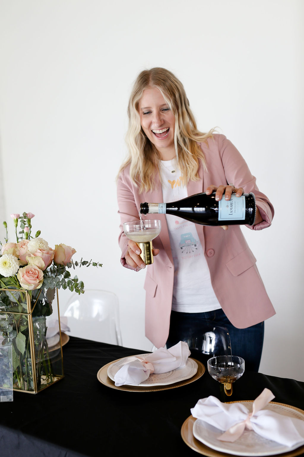 Woman pouring champagne into a glass at a table with flowers and place settings, smiling, in a bright room.