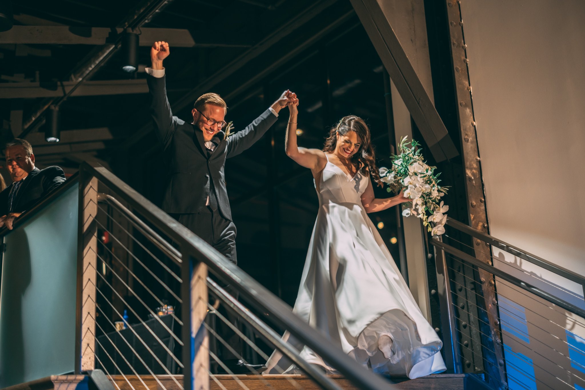 A bride and groom dance on a balcony, holding hands, with the bride holding a bouquet of flowers, in a dimly lit setting.