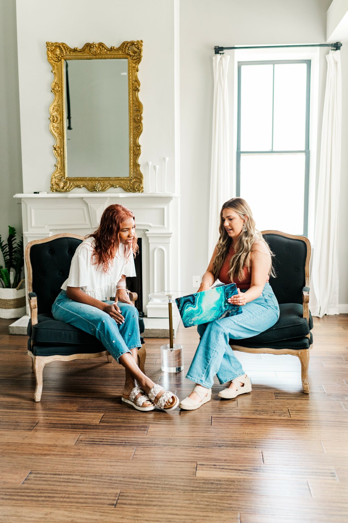 Two women sit on black and white chairs in a bright room with a large window, a gold-framed mirror, and a white fireplace.