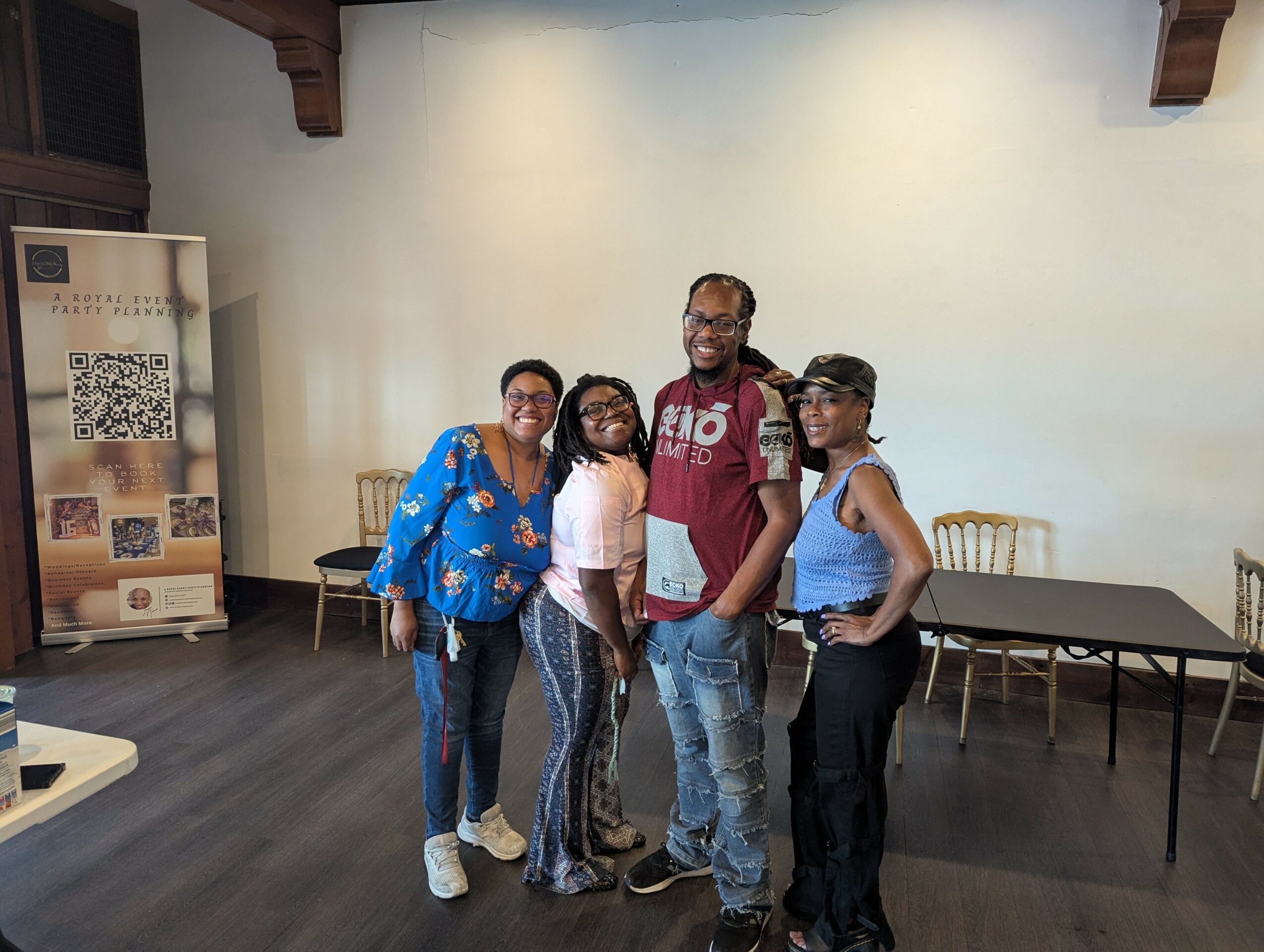 Group of five people standing indoors, smiling, with chairs and a table in the background.