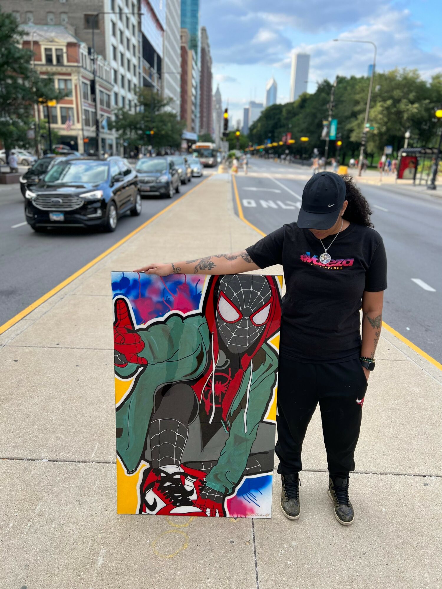 Person in black cap and shirt stands on city sidewalk next to colorful Spider-Man artwork, with cars and buildings in background.