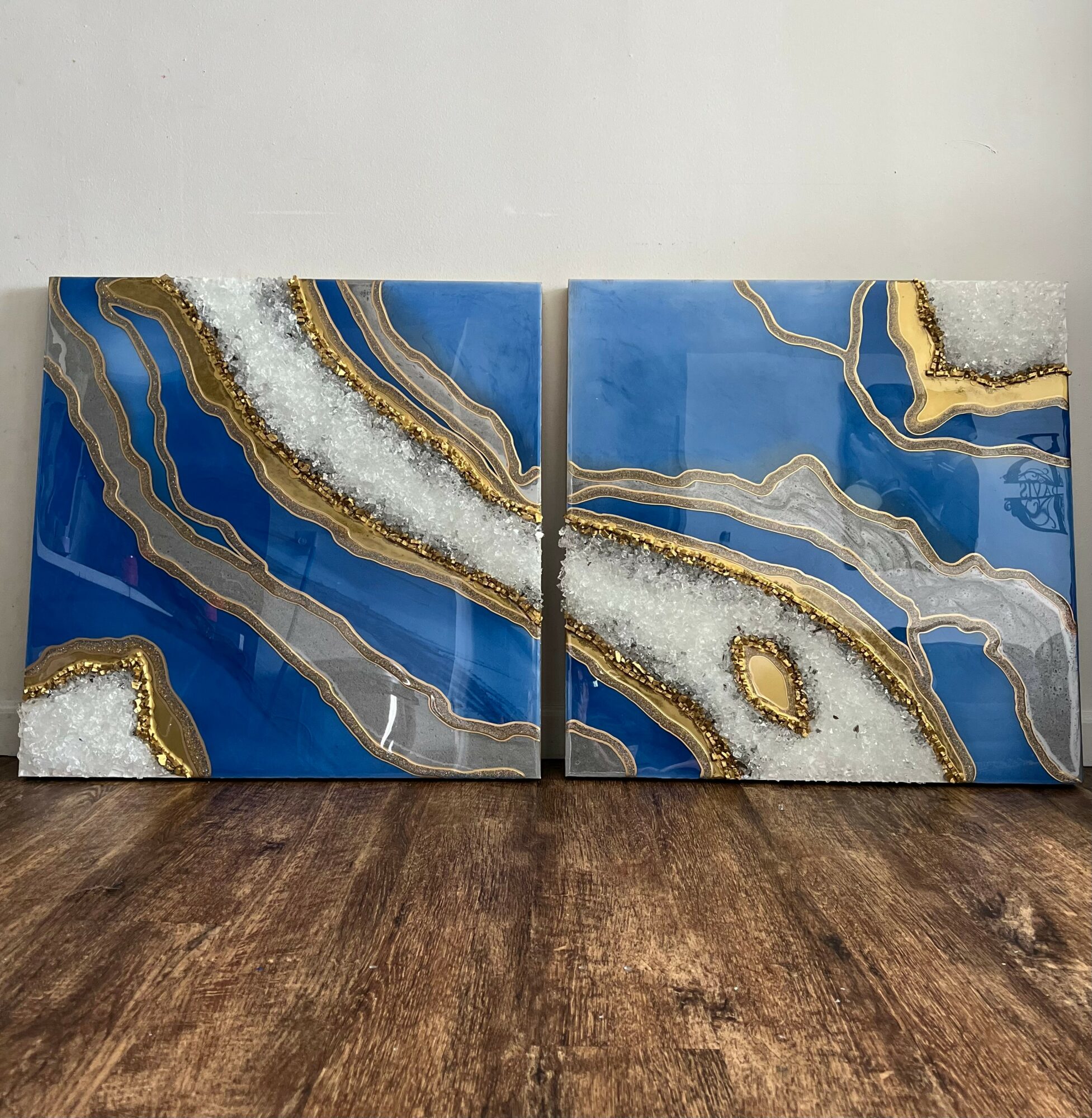 Two blue and gold geode-style art pieces with white crystalline accents, placed on a wooden floor against a plain wall.
