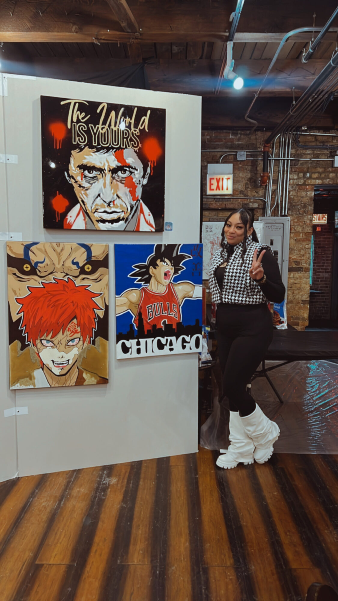 Woman standing next to four colorful anime-style posters on a white display, in a brick-walled room with wooden floor.