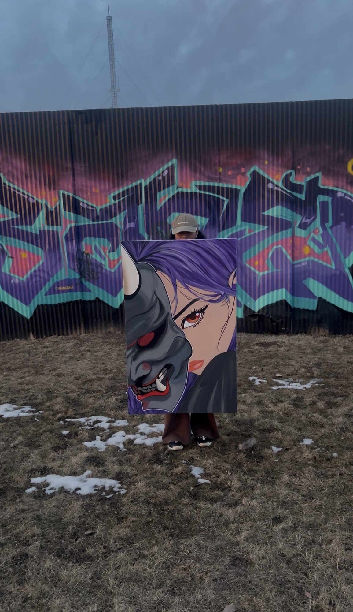Person holding a large graffiti-style portrait of a woman with purple hair and a black mask, standing outdoors with a colorful graffiti wall behind.