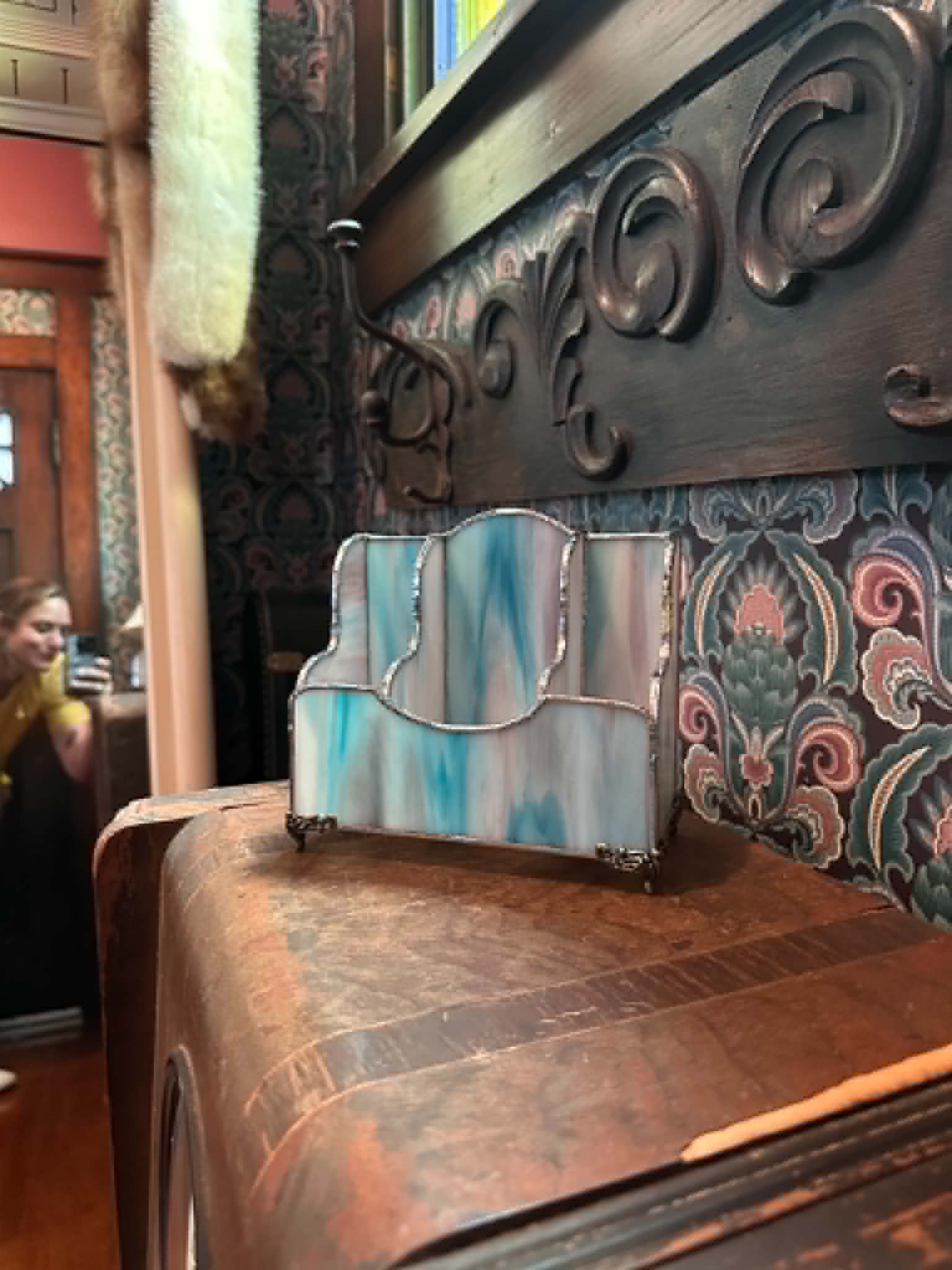 Decorative metal object on a wooden surface with patterned wallpaper in background, person taking a photo reflected in mirror, 20 words